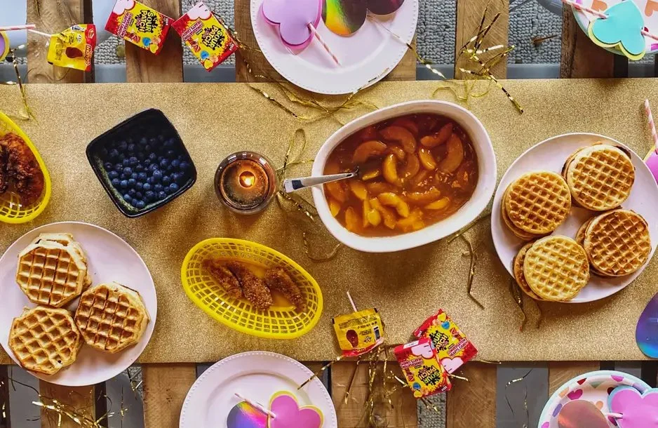 Party table set with waffles, fruit-filled compote, blueberries, candy, and decorative heart-shaped items.