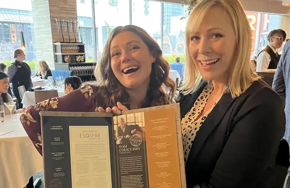 Two women smiling in a restaurant, holding a menu or award, with other diners and large windows in the background.
