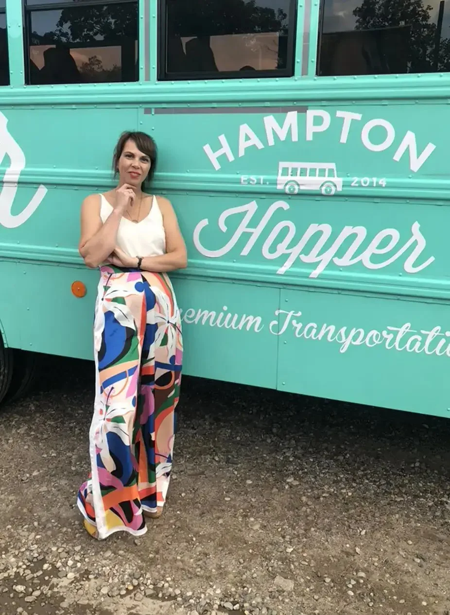 A woman standing next to a teal-colored bus labeled 'Hampton Hopper' with an illustration of a bus, promoting premium transportation, on a gravel ground outdoors.