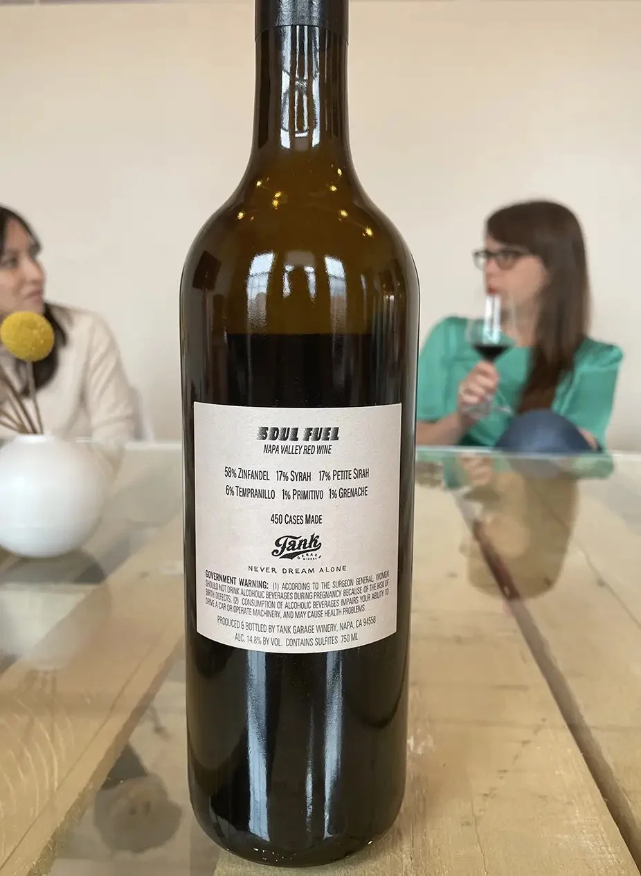 A bottle of red wine labeled 'Soul Fuel,' from Napa Valley, with a group of people in the background, two women clearly visible, one drinking red wine and the other looking at her, in a casual indoor setting.