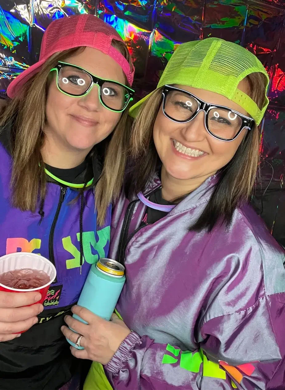 Two women smiling, wearing colorful neon hats, glasses, and shiny jackets, holding drinks, with a vibrant, holographic background.