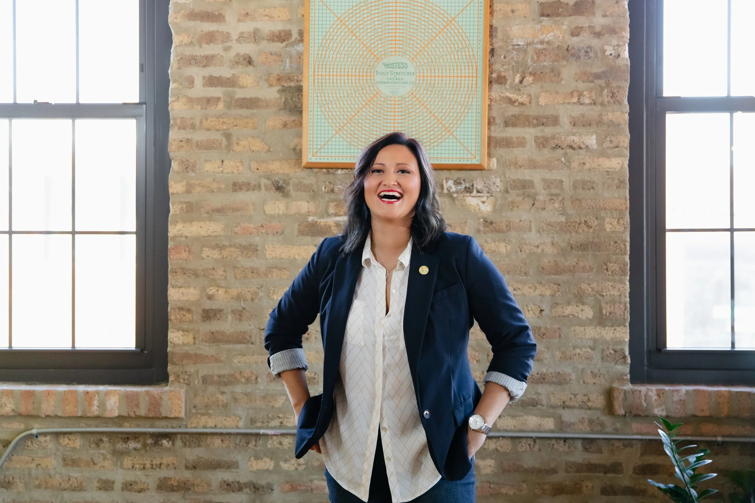 Smiling woman with shoulder-length dark hair in a navy blazer and white shirt standing with hands on hips in front of a brick wall and large windows, with a large sewing or drafting pattern on the wall behind her.