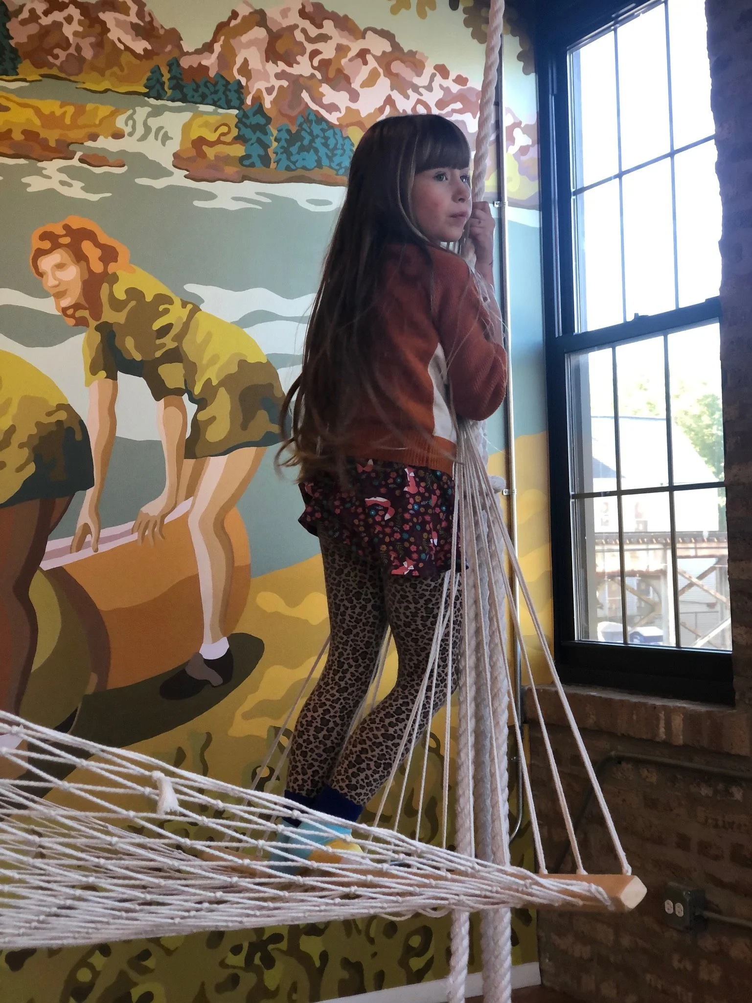 A young girl with long brown hair, wearing a brown jacket, animal print leggings, and colorful socks, standing on a small wooden and rope platform near a window. Behind her is a mural depicting children playing or working with large logs in a nature 