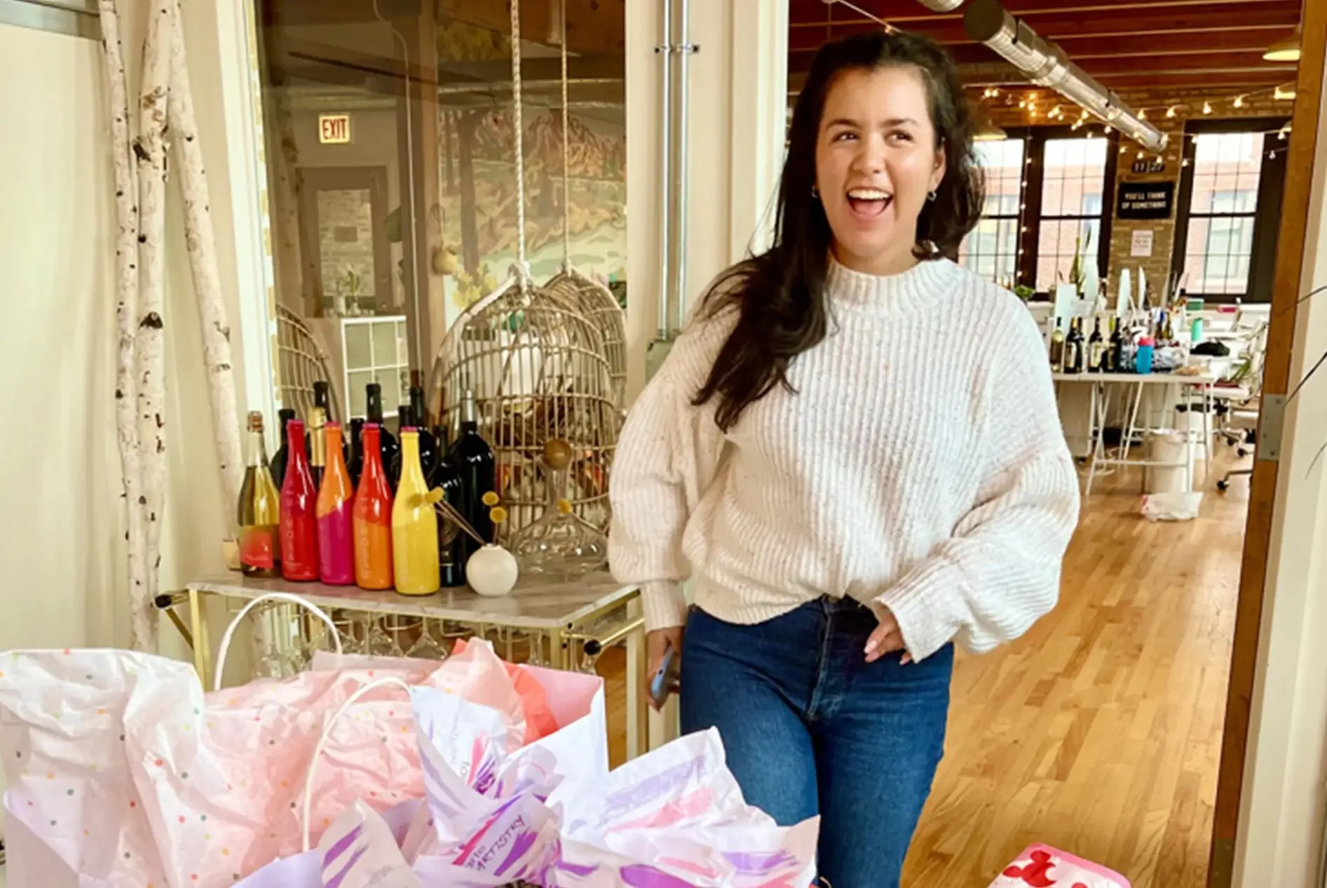 A woman smiling and posing, standing in a bright, cozy room with wooden floors and colorful decorations. There are gift bags in the foreground, and bottles and decorative items on a small table behind her.