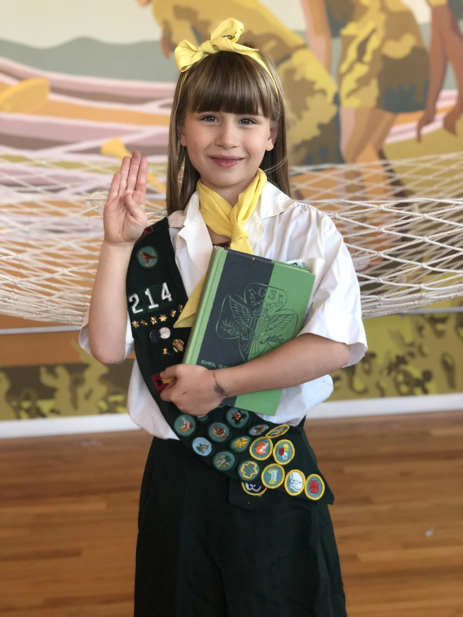 Girl scout girl in uniform, wearing a yellow neckerchief and a headband, holding a folder and making the scout sign with her right hand, standing indoors with a mural in the background.