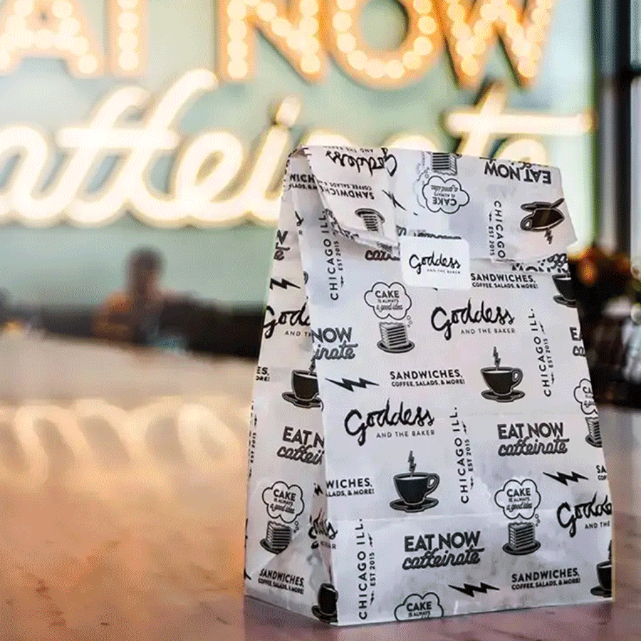 A paper bag with various bakery and cafe-themed graphics and slogans, placed on a wooden table inside a cafe.