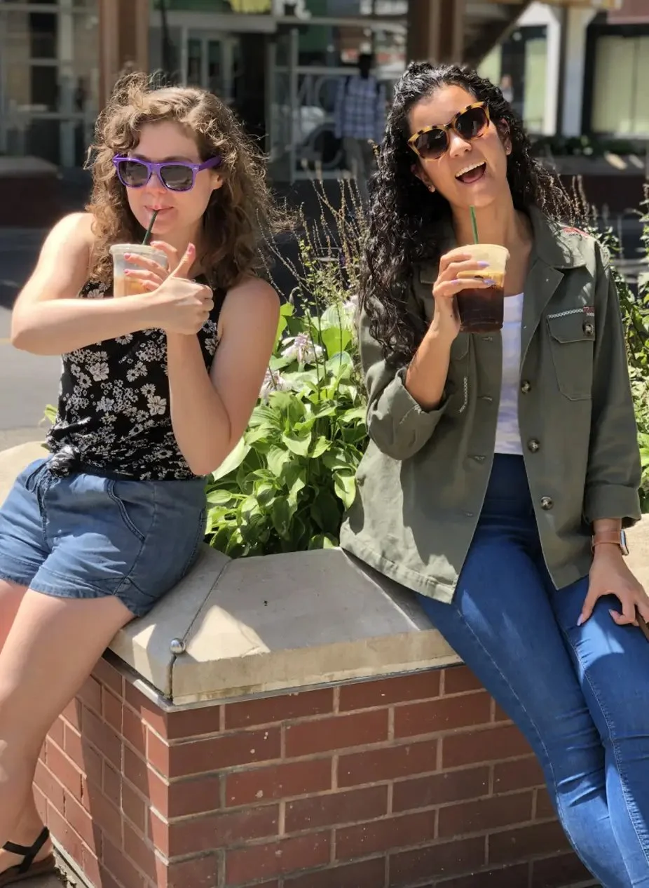 Two women sitting on a brick ledge outdoors, drinking iced coffee or frappuccinos, both wearing sunglasses and smiling.