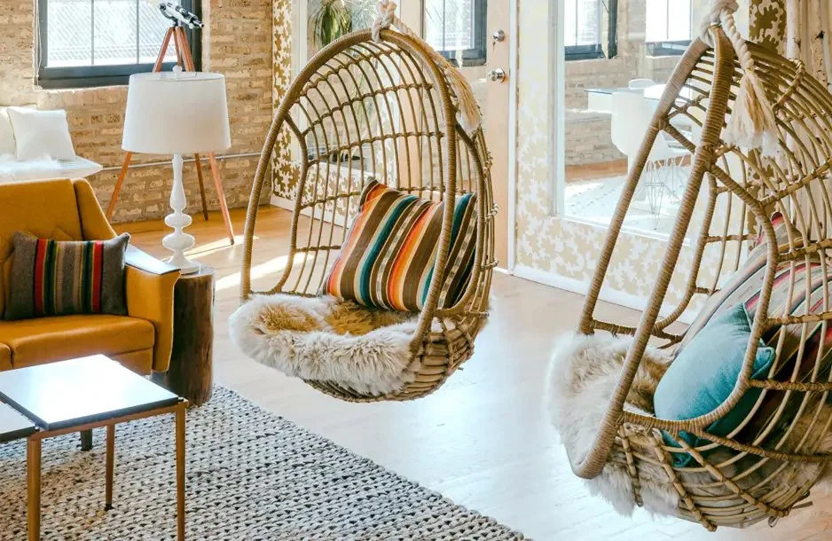 Living room with two hanging rattan egg chairs with cushions and fur throws, a mustard-colored sofa with striped pillows, a white lamp on a wooden tripod table, a black and white patterned rug, and large windows with brick walls and a glass door lead