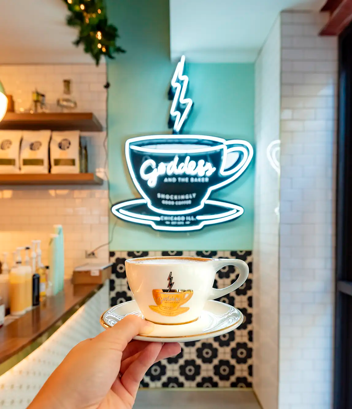 A hand holding a coffee cup on a saucer in front of a neon sign of a coffee cup with lightning bolt above and text 'Goddess and the Baker'. The background features a decorated cafe interior with white tiled walls, shelves, and a patterned wall.