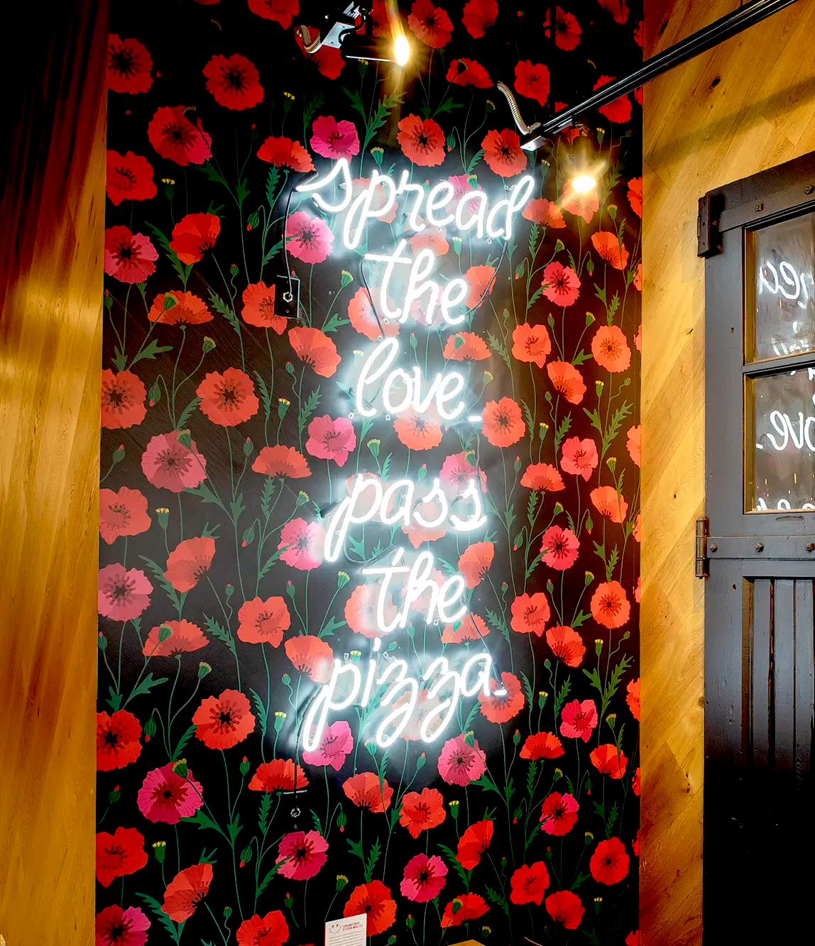 A neon sign with the words "Spread the love pass the pizza" mounted on a floral wallpaper background with red and pink flowers and green leaves, inside a room with wooden walls and a black door.