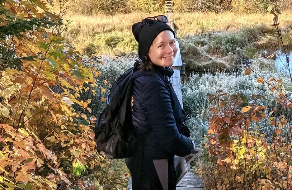 A woman in outdoor clothing, including a black beanie and backpack, standing on a wooden bridge surrounded by autumn foliage and a frosty pond.