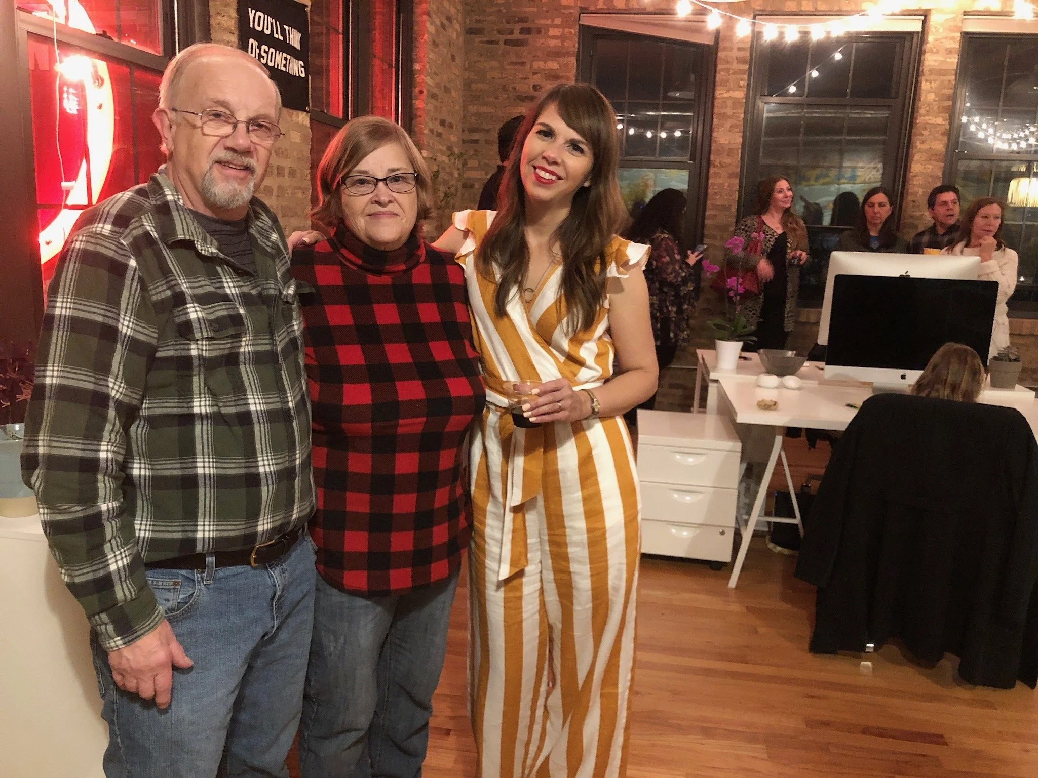 Three people standing together at a social gathering in a cozy, warmly lit room with brick walls and large windows. The man on the left has white hair, glasses, a beard, and is wearing a plaid shirt. The woman in the middle has short hair, glasses, a