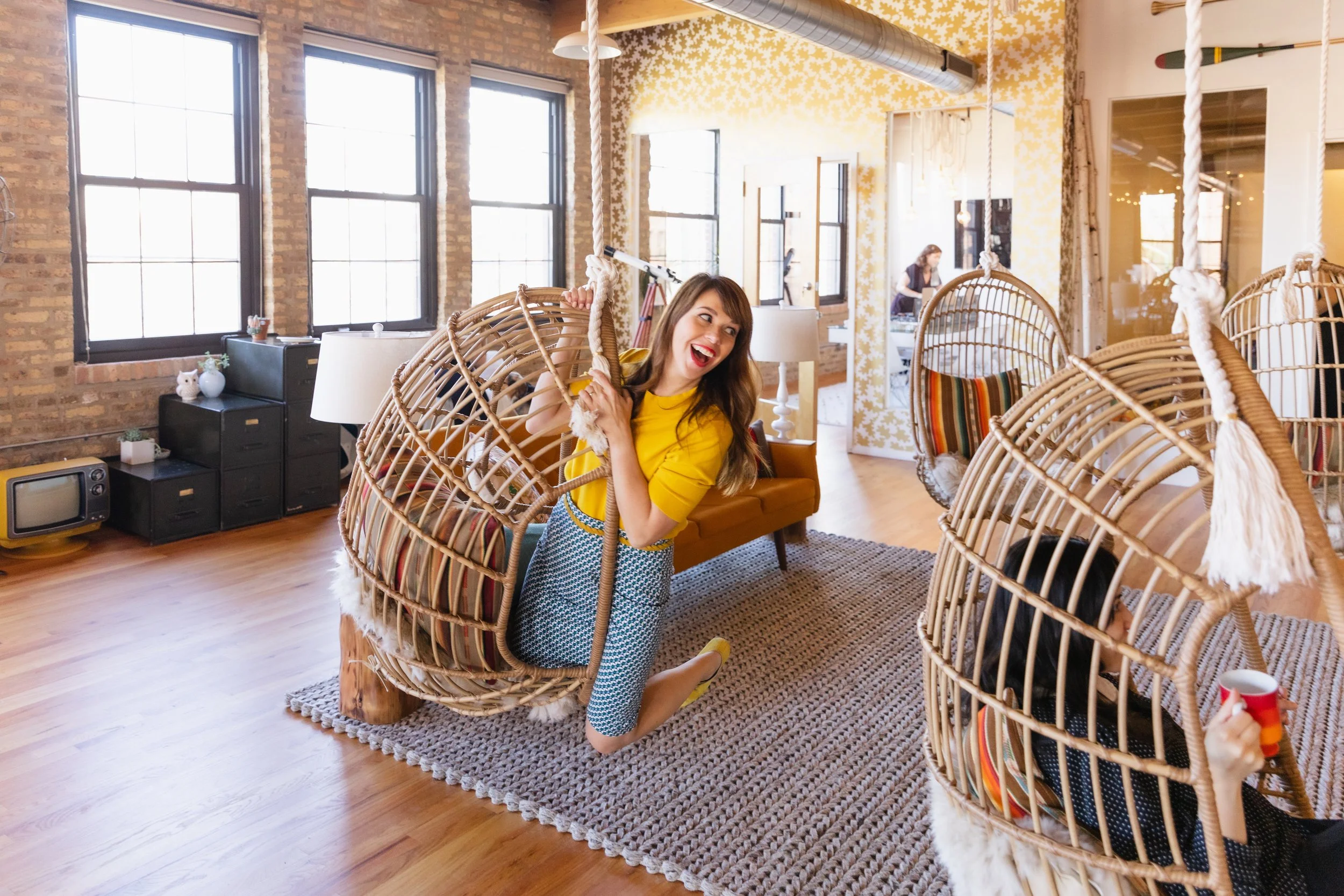 A woman in a yellow top and checkered pants sitting inside a hanging wicker chair, smiling and playing with another hanging wicker chair with a woman holding a mug, in a cozy, modern living room with large windows, wooden floors, and eclectic decor.