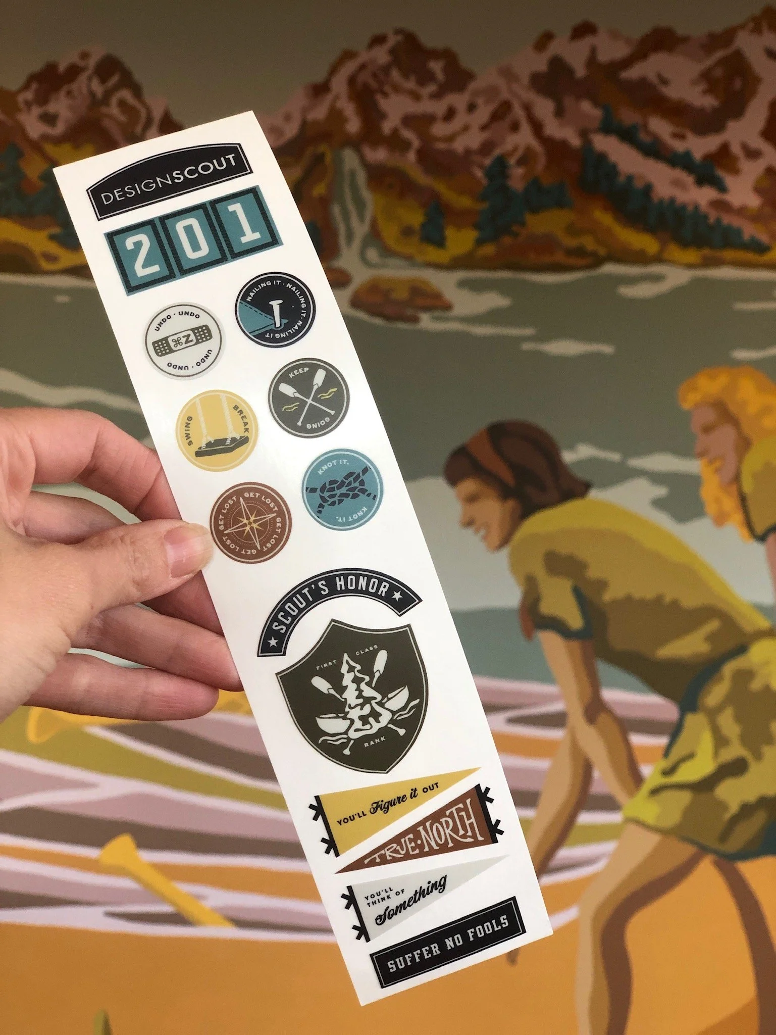 A hand holding a sheet of Scout badges in front of a colorful mural of two children in yellow shirts by a body of water with mountains in the background.