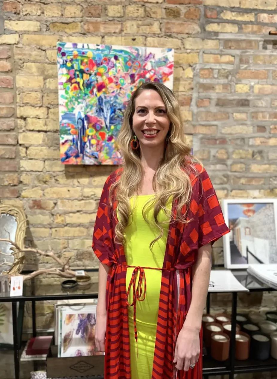 A woman with long blonde hair, wearing a bright yellow dress and red and black patterned kimono, stands in an art gallery with brick walls. She is smiling and posing in front of colorful abstract artwork.