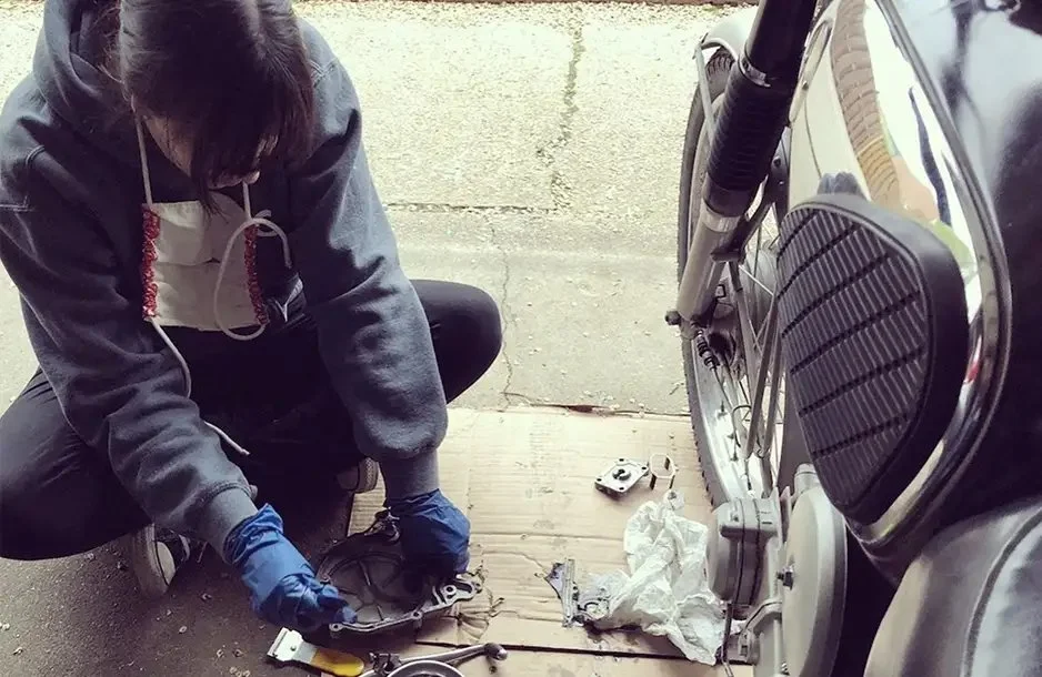 A person wearing a gray hoodie, black pants, and blue gloves is working on a motorcycle in a garage or driveway. They are crouched down and appear to be repairing or inspecting part of the motorcycle. The motorcycle is black and silver and is parked 