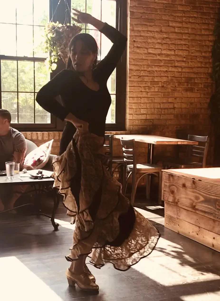 A woman dancing indoors near a window with sunlight, wearing a black top and flowing, ruffled skirt, on high dance heels, with a man sitting at a table behind her.