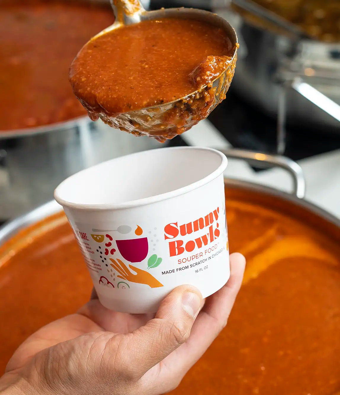 A metal ladle filled with tomato soup being poured into a white paper bowl labeled 'Sunny Bowls Souper Food' with colorful graphics.