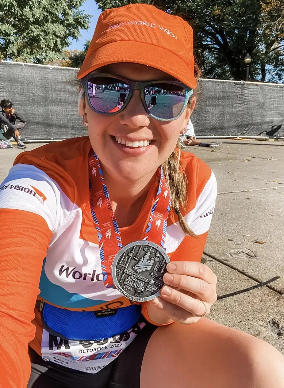 A woman wearing an orange cap, sunglasses, and a multicolored race shirt, smiling and displaying a medal from the Chicago Marathon on October 9, 2022, at an outdoor event.