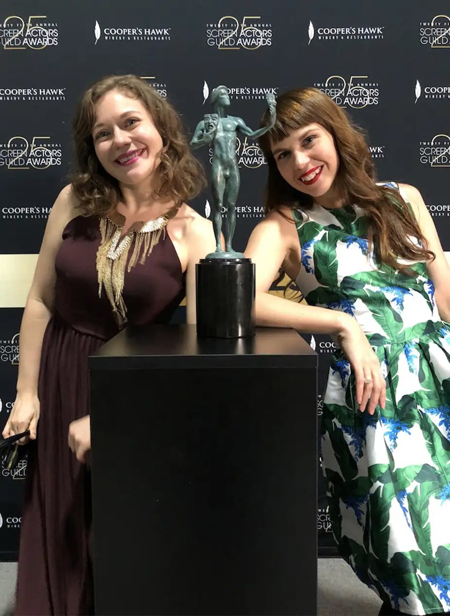 Two women smiling at an event standing behind a black pedestal with a bronze trophy on top. The trophy features a tall, slender figure holding an object in one hand and covering their face with the other. The background has a step-and-repeat banner w