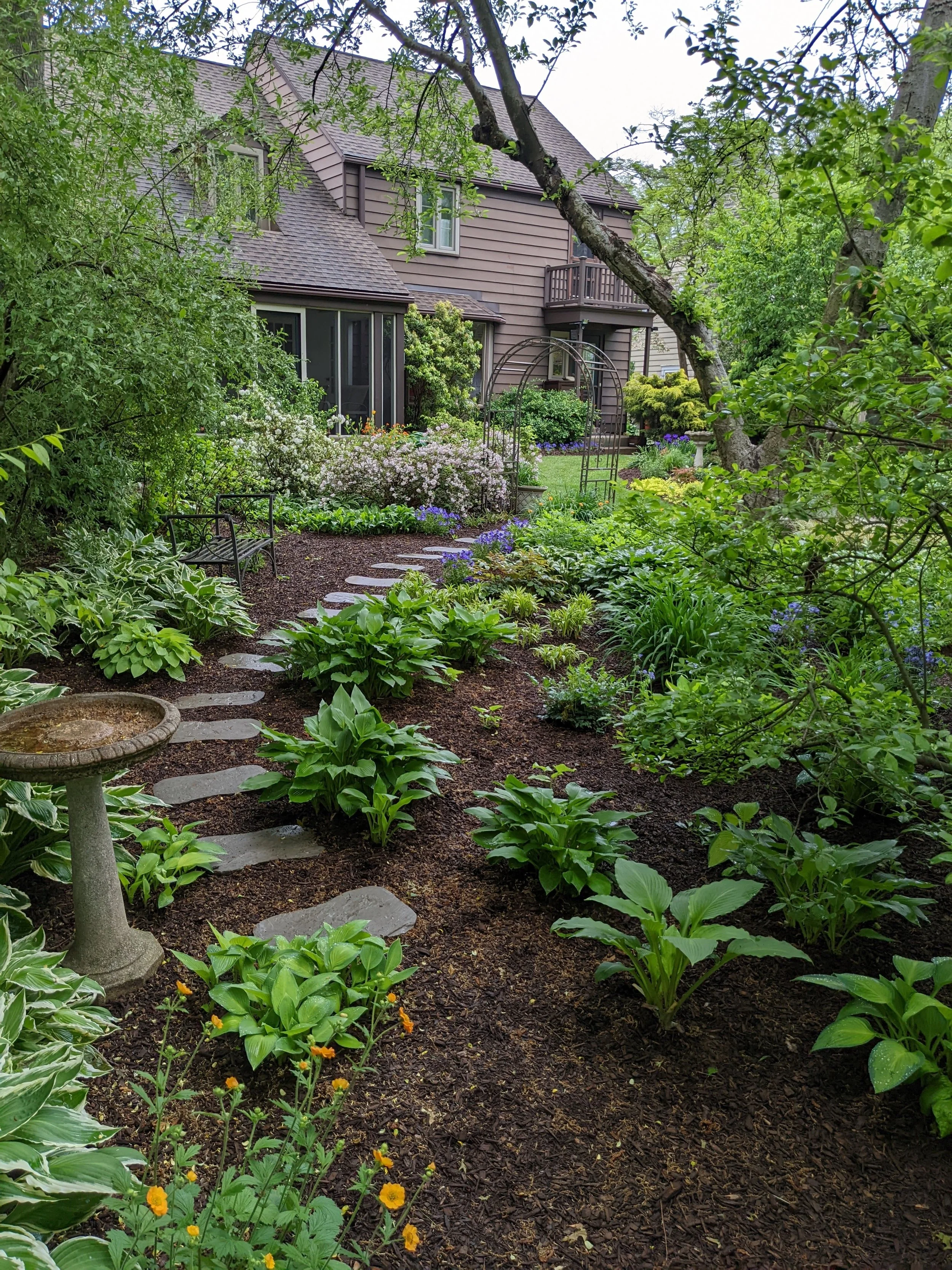 A lush garden with various plants, flower beds, a stone pathway, garden bench, birdbath, and a large house with a balcony in the background.
