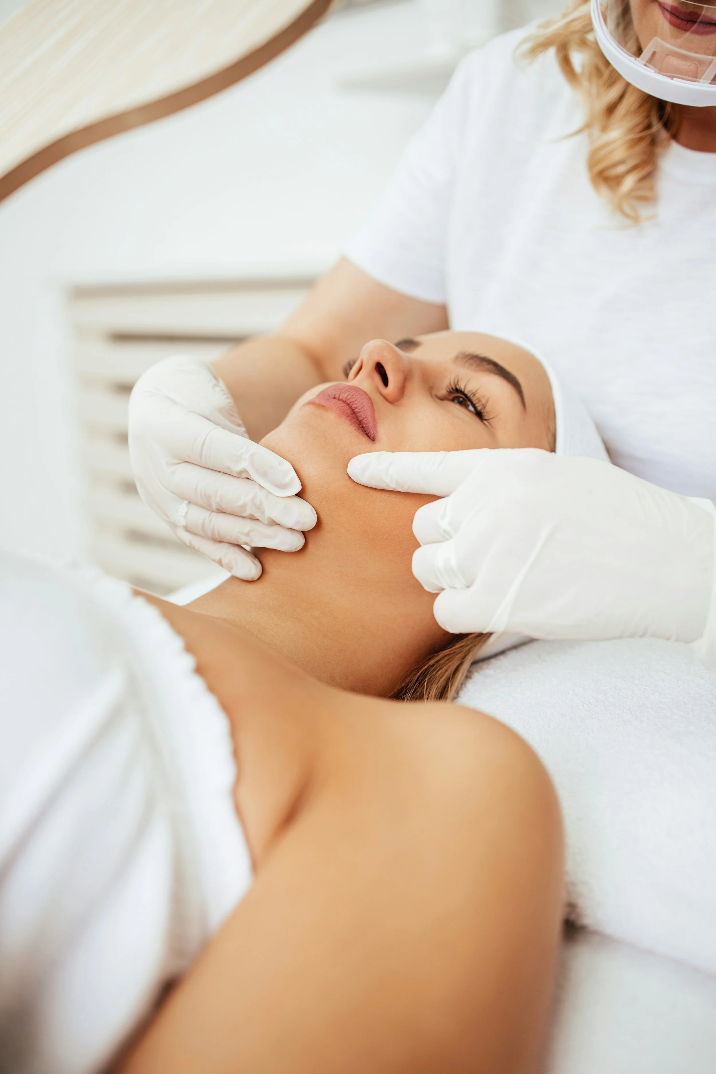 A woman receiving a facial treatment at a spa, lying on a treatment bed with her eyes closed, while a professional in white gloves gently holds her face.