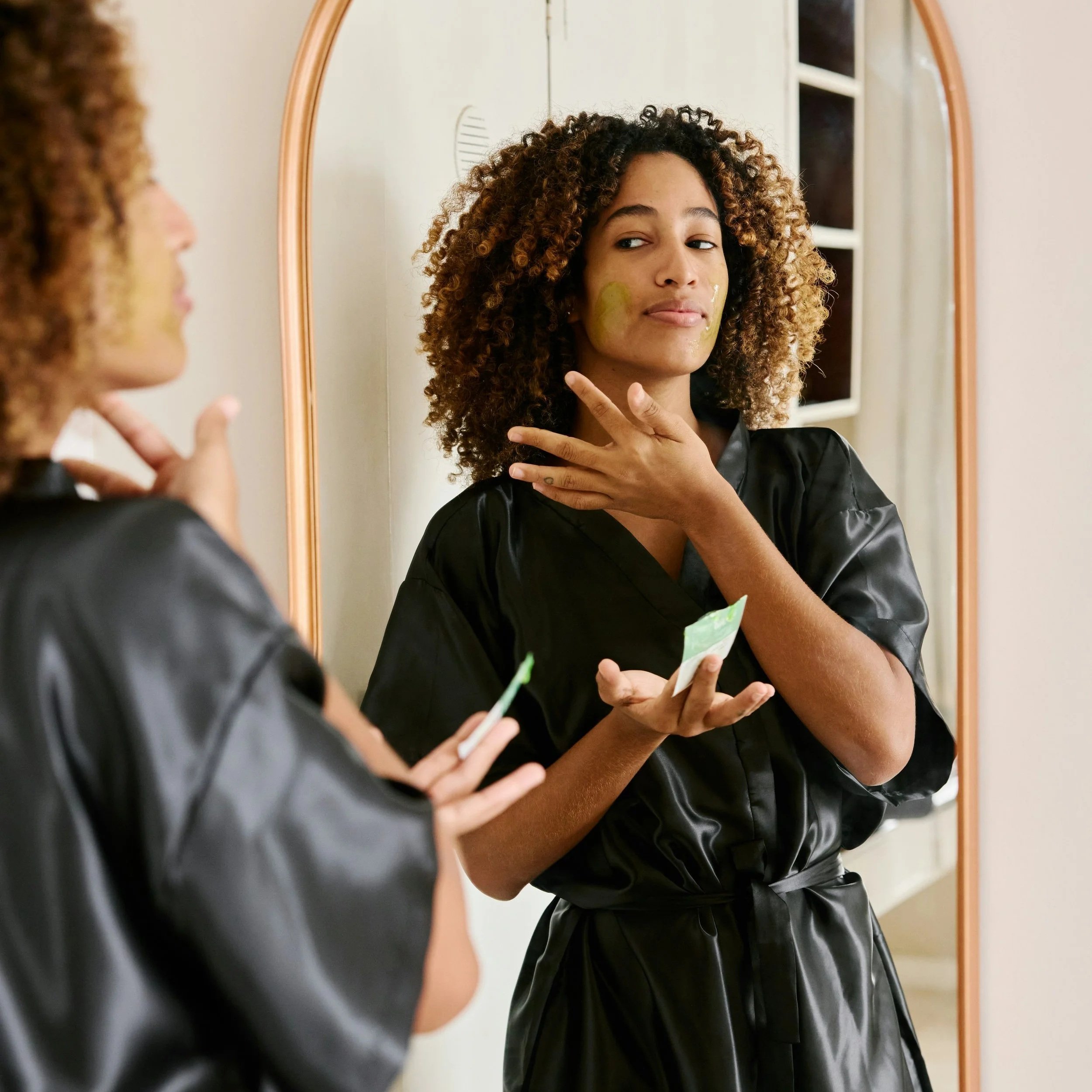 A woman with curly hair is looking at herself in a mirror while applying skincare or cosmetic products on her face, holding a tube or packet in one hand.