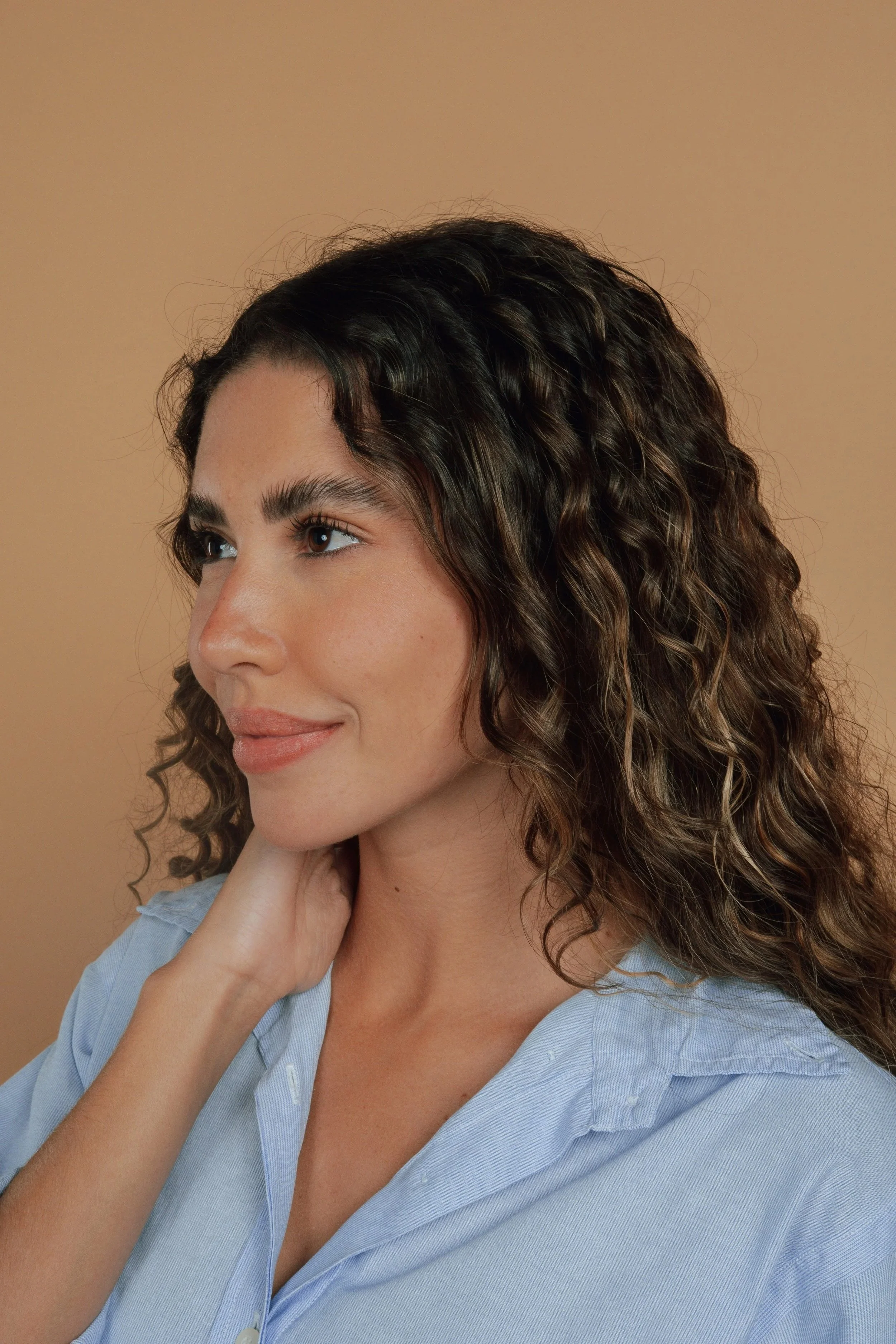 Close-up of a woman with long, curly brown hair, wearing a light blue shirt, touching her neck, against a warm beige background.