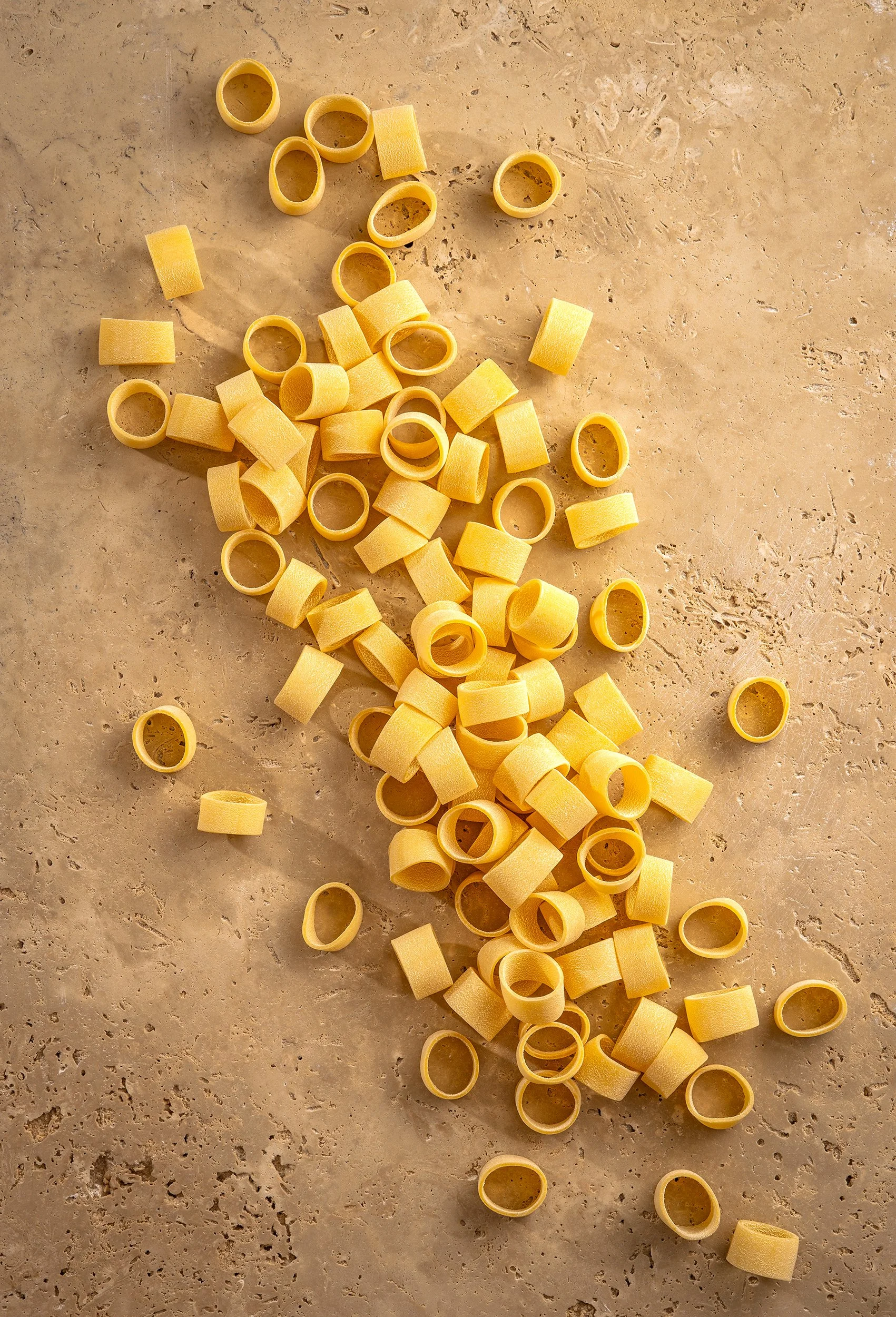 Scattered yellow pasta rigatoni and orecchiette on a beige marble surface.