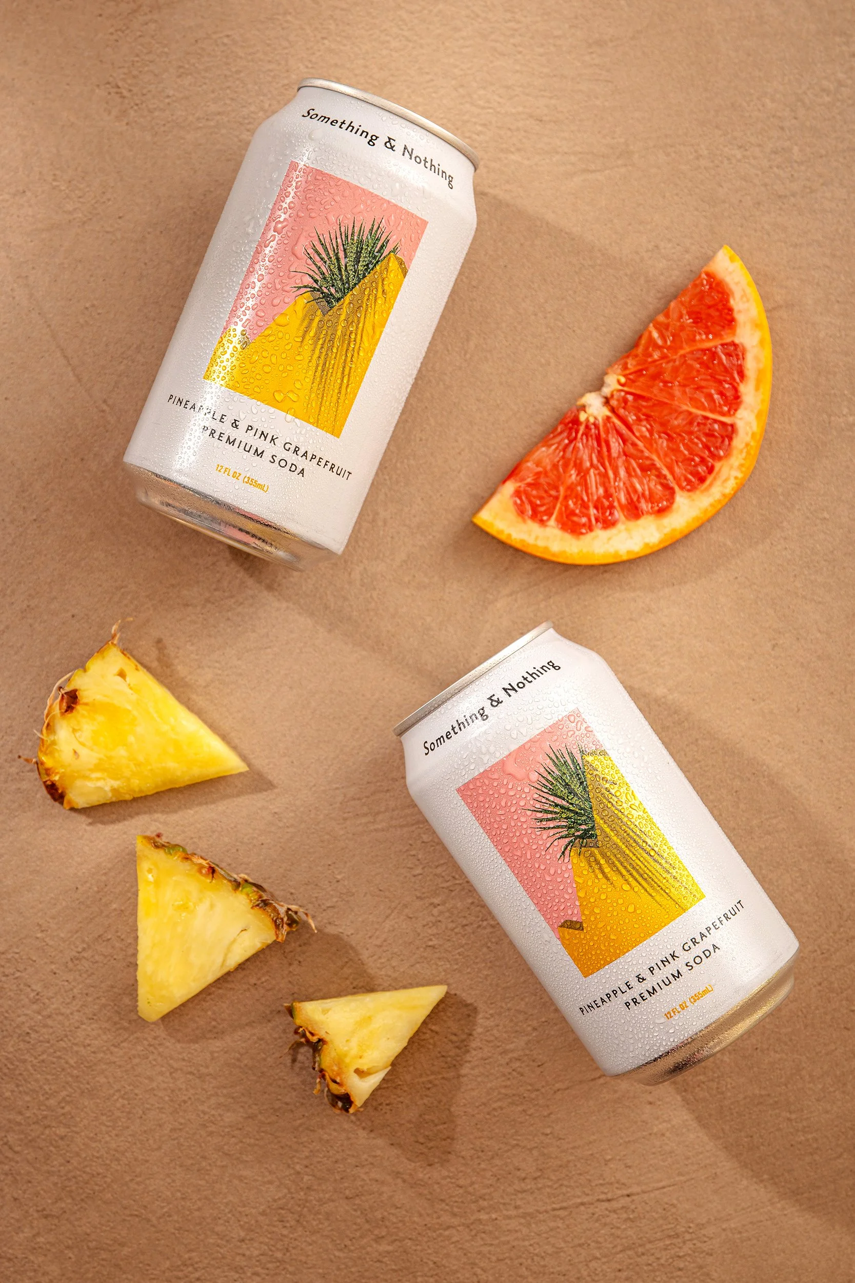 A can of pineapple and pink grapefruit soda with condensation droplets on the exterior, placed on a tan surface next to sliced pineapple and a grapefruit wedge.