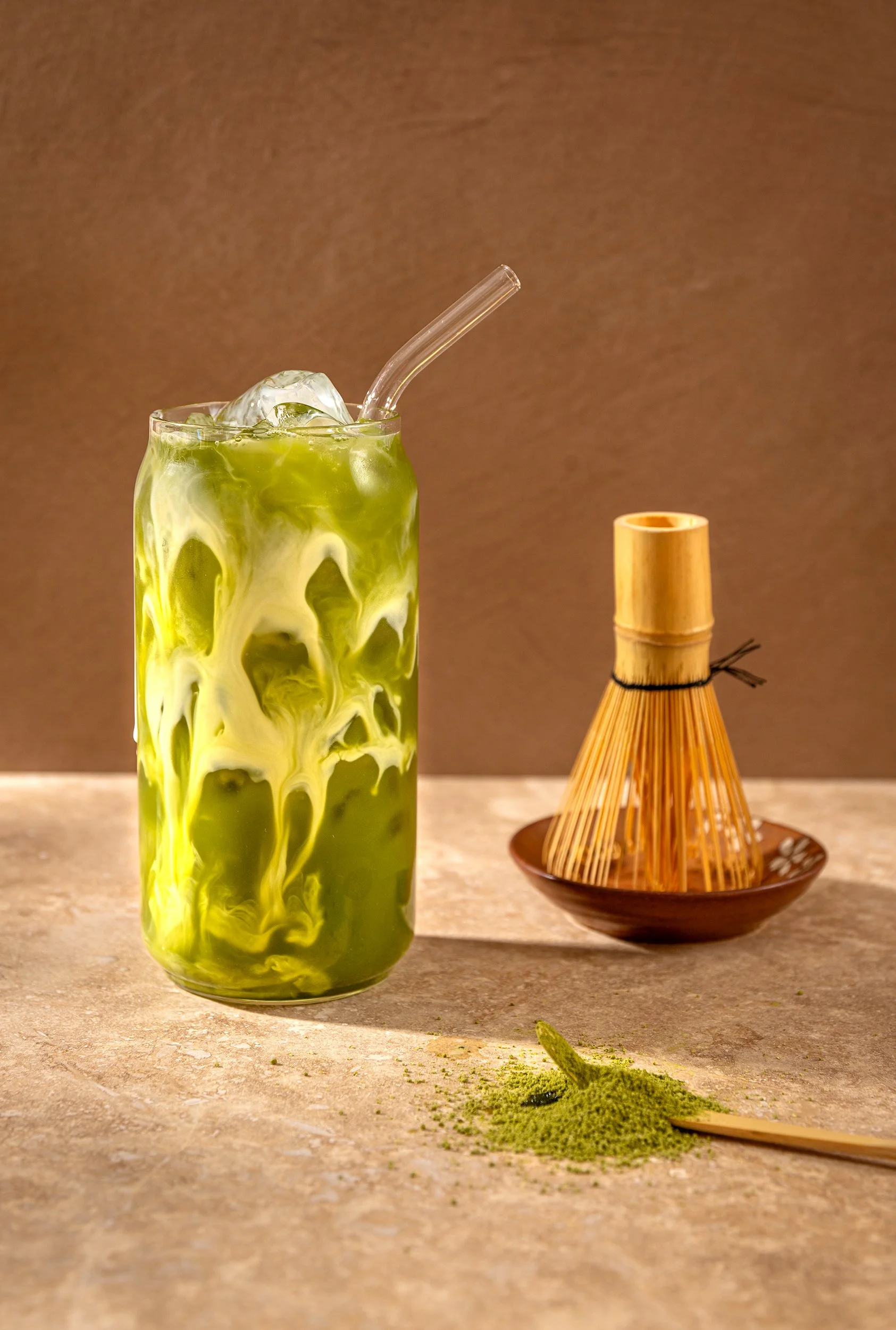 A tall glass of green matcha latte with ice cubes and a clear straw, on a beige surface. To the right, a bamboo tea strainer and a small dish with green matcha powder, some of which is spilled on the surface.