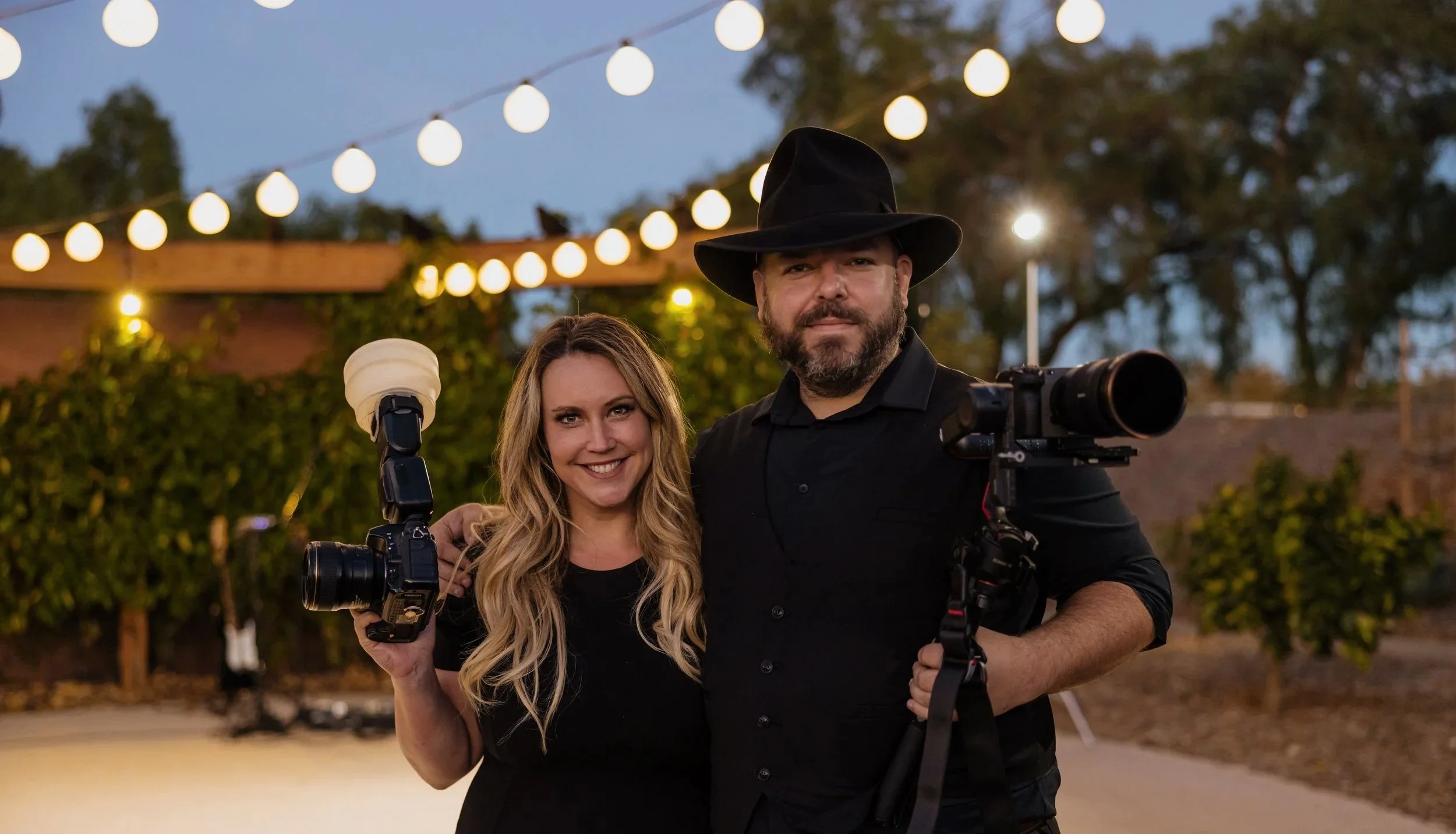 A smiling woman holding a camera and a smiling man with a camera and a large lens, standing outdoors during the evening under string lights with trees in the background.