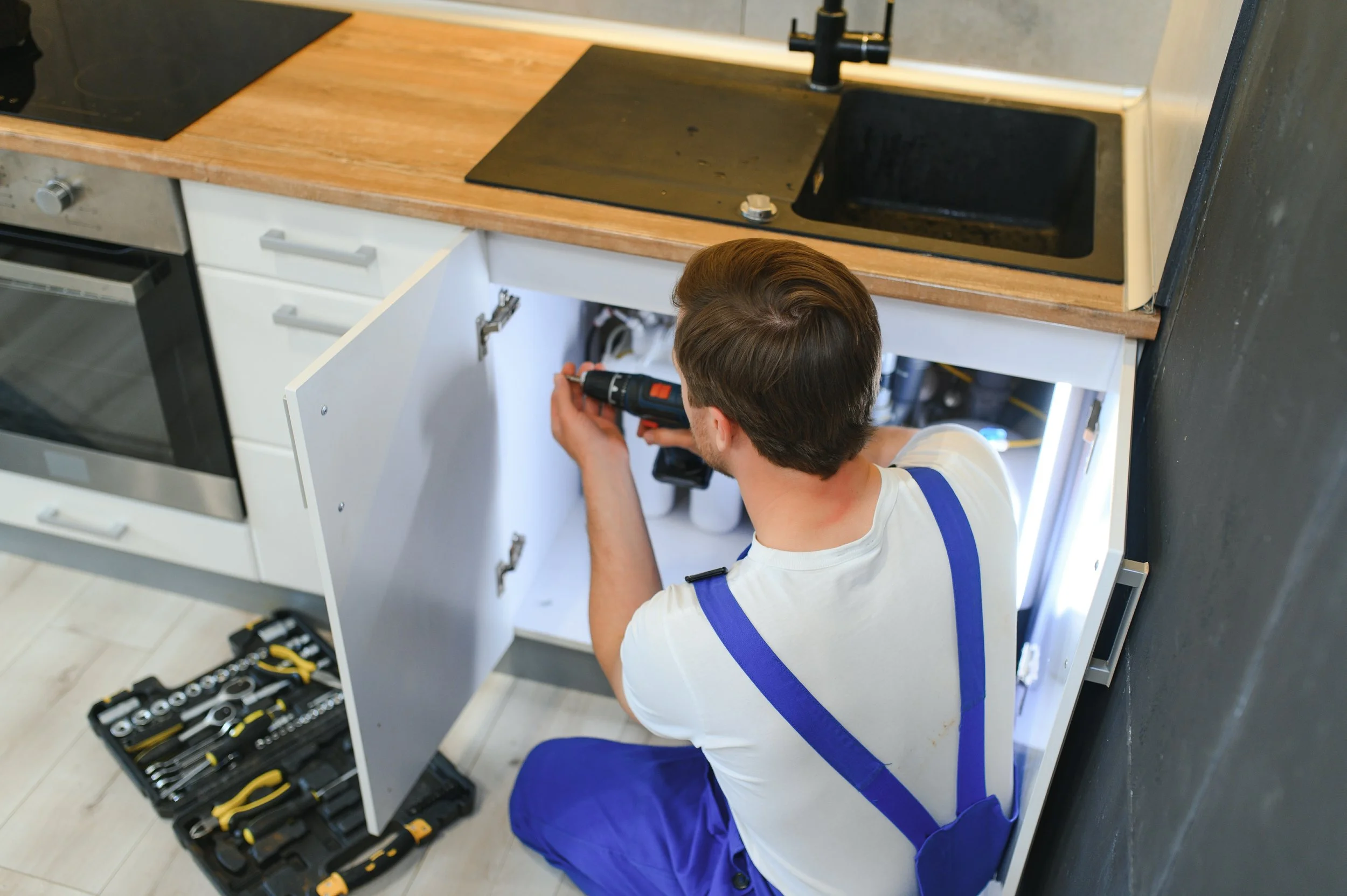 A man wearing a white t-shirt and blue overalls repairing a kitchen cabinet with a cordless drill. There is an open toolbox with various tools on the floor nearby.