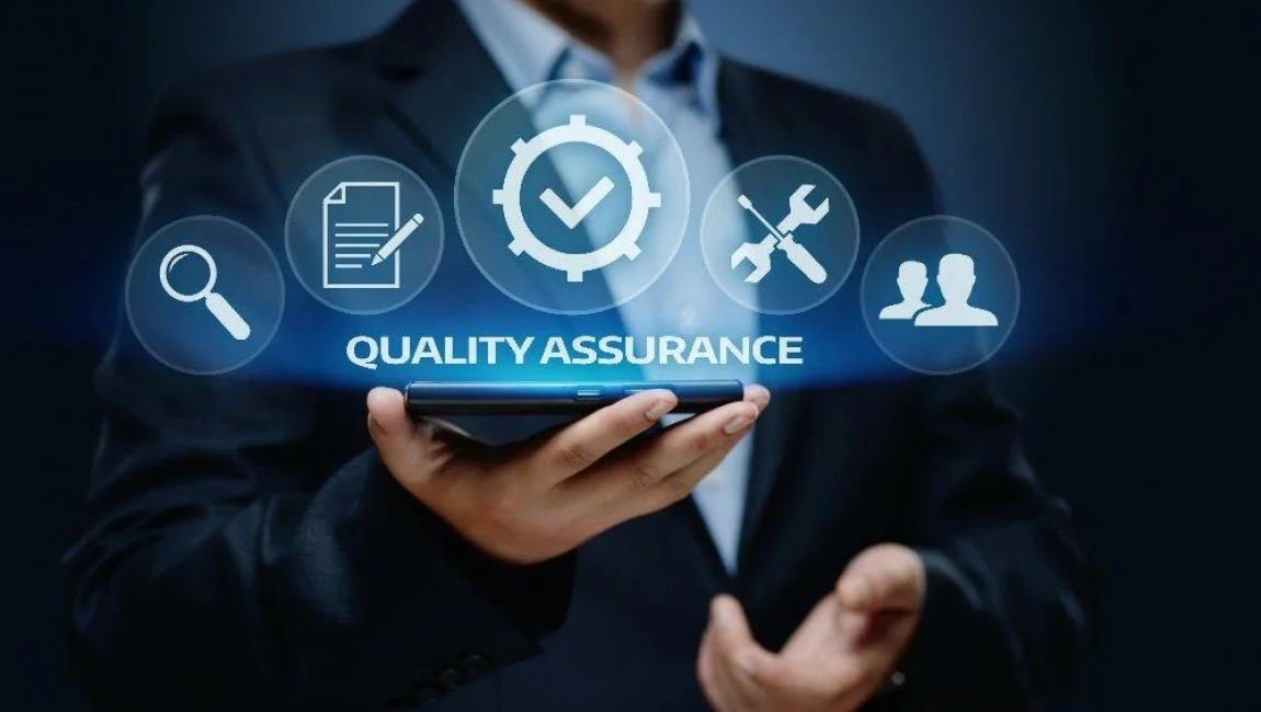 A person in a business suit holding a smartphone displaying a digital graphic with icons representing quality assurance, including checkmark, magnifying glass, document, screwdriver and wrench, and two people, with the words "QUALITY ASSURANCE" underneath.
