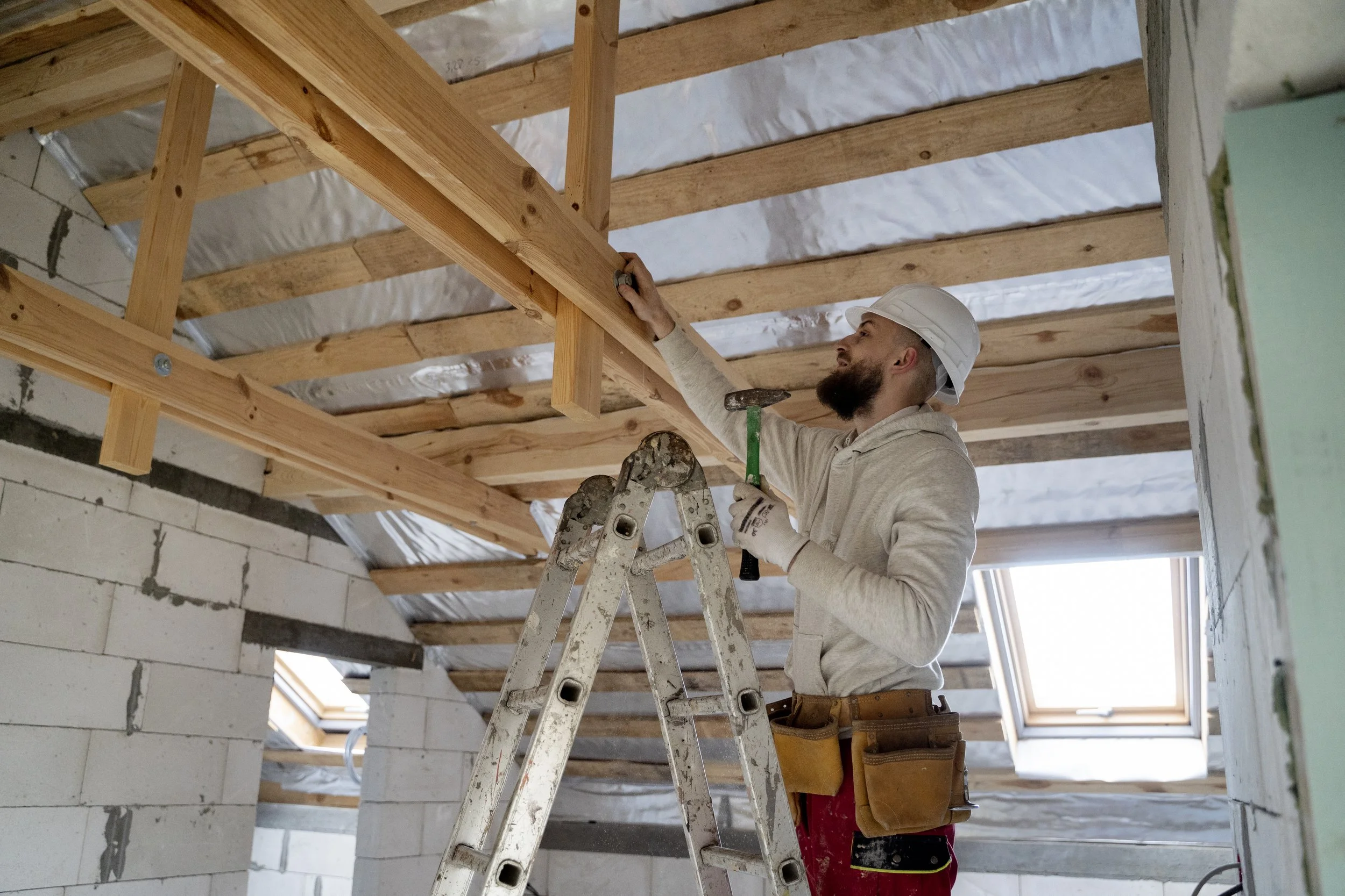 A construction worker standing on a ladder installing or inspecting wooden beams in a building under construction. The worker is wearing a white hard hat, gloves, a beige hoodie, red pants, and a tool belt.