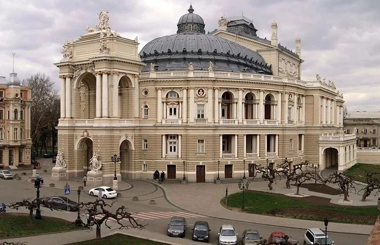 A grand, historic European-style building with ornate architectural details, a large dome, and multiple arches, situated in an urban area with parked cars and leafless trees.
