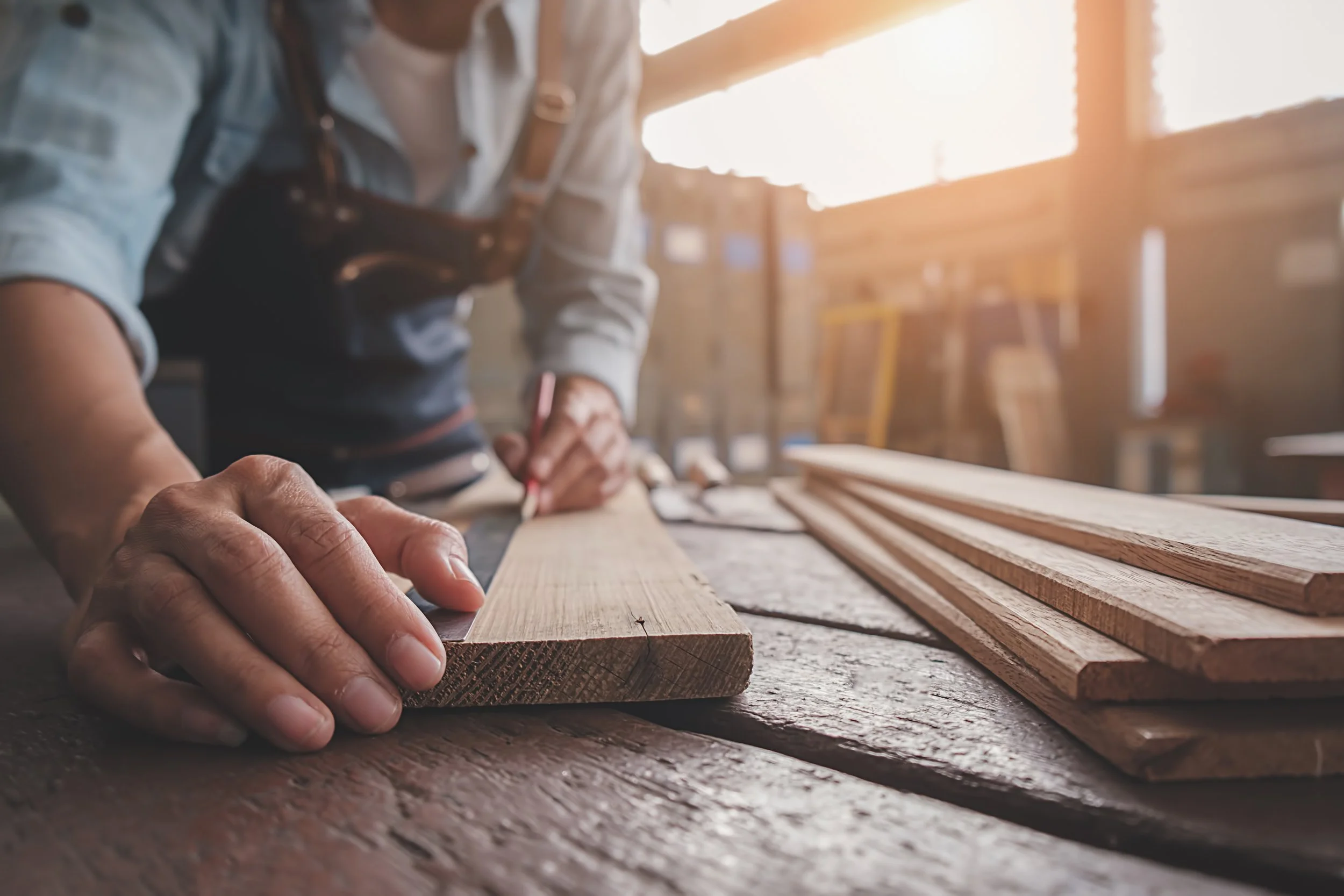 Person measuring and marking a piece of wood in a woodworking shop with sunlight streaming through windows.