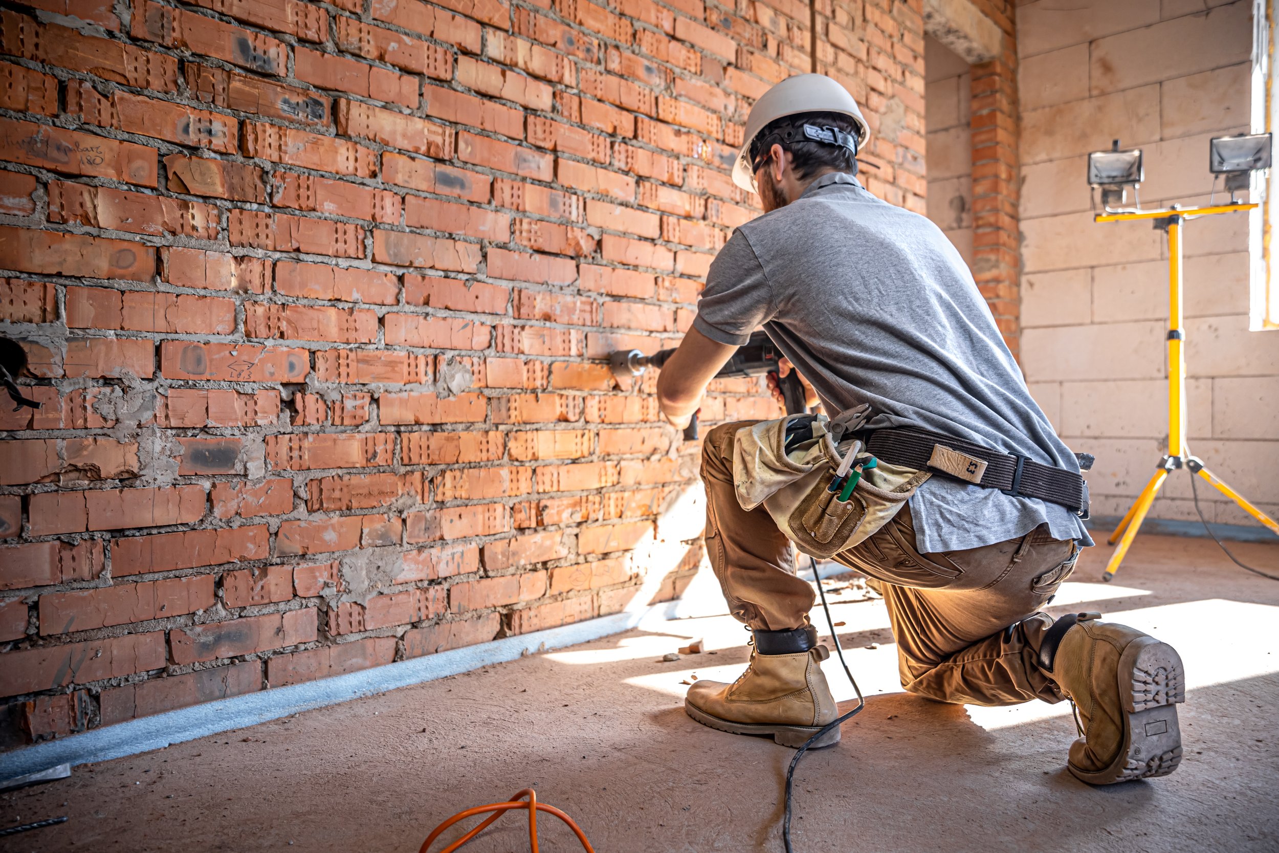 A construction worker wearing a helmet, gray shirt, brown pants, and tan boots is kneeling on the floor while using a power drill on a brick wall. There is a yellow scaffolding and a light stand in the background, with sunlight coming through a window.