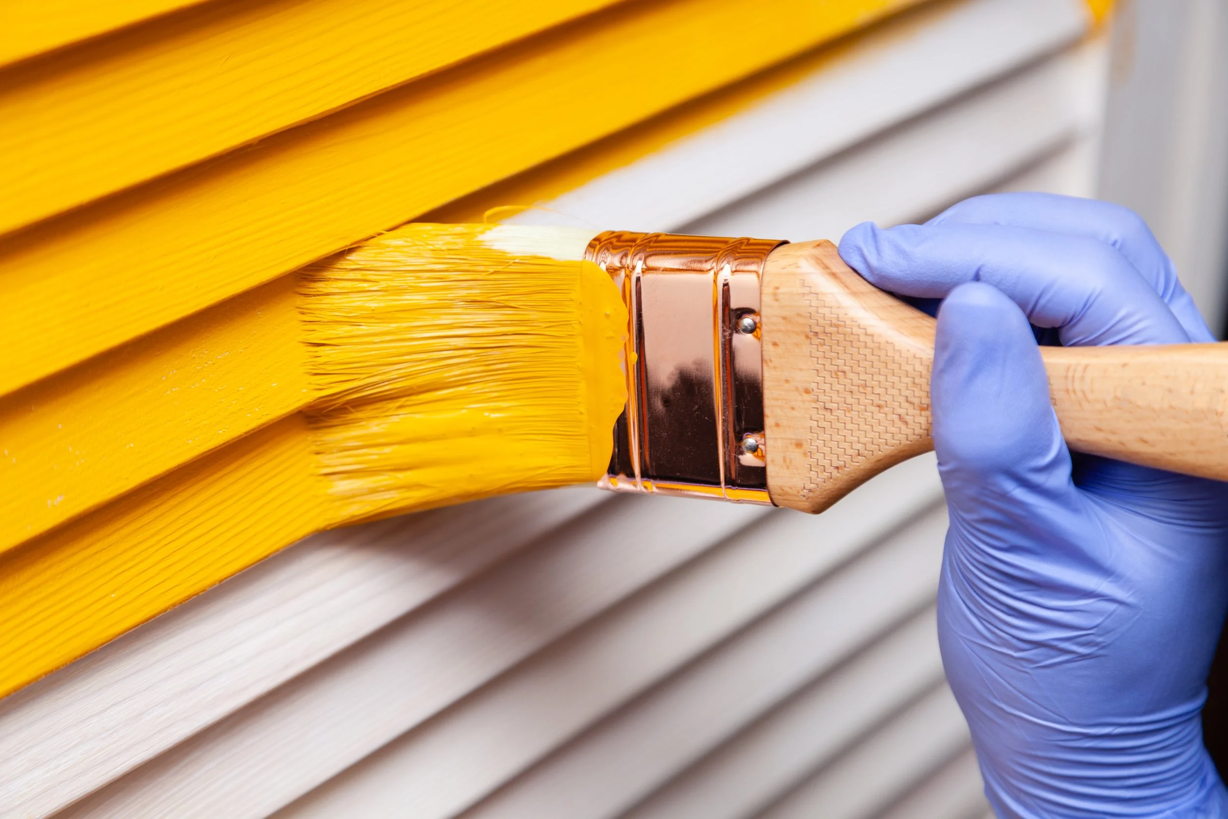 Person wearing blue gloves painting yellow horizontal wooden slats with a paintbrush.