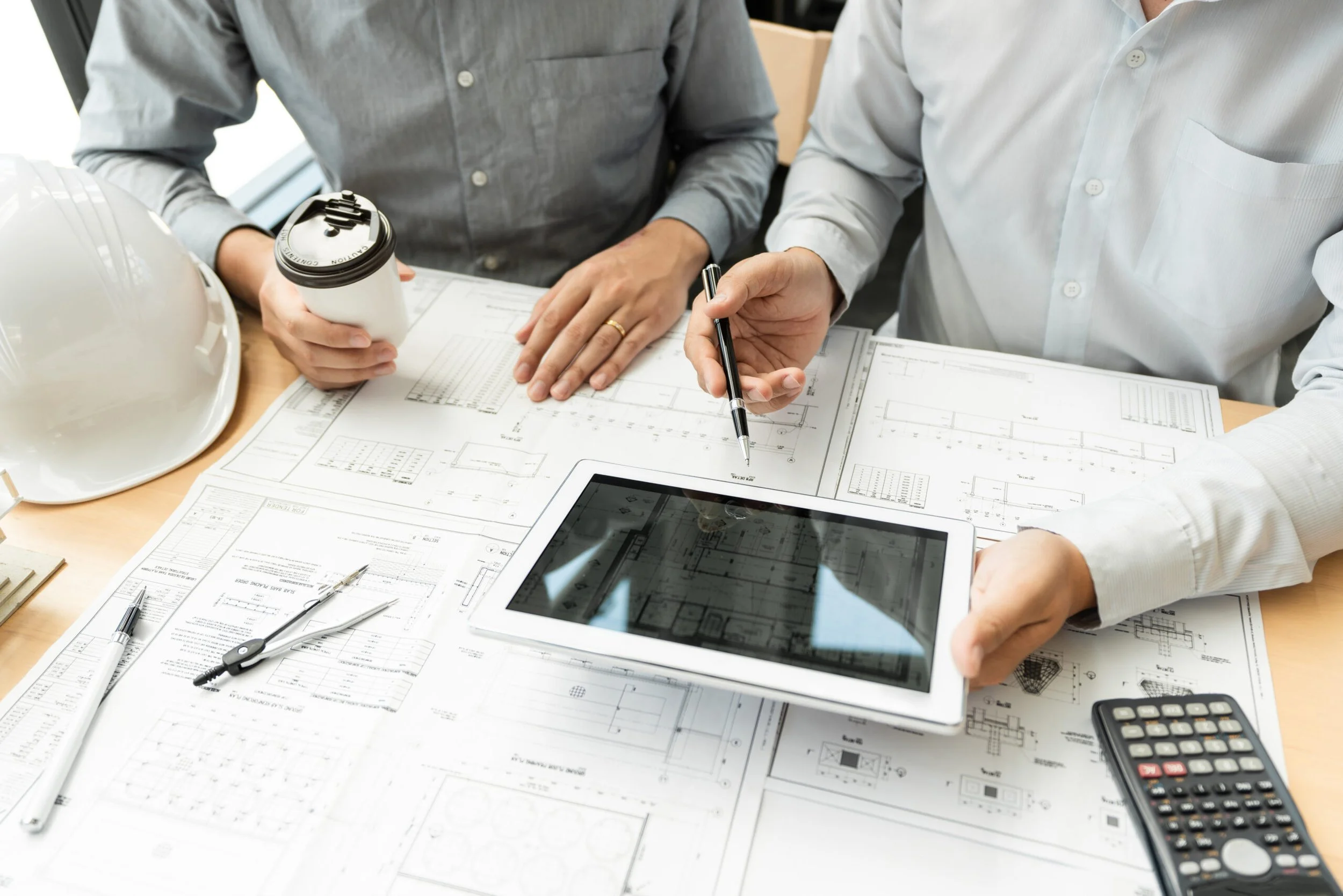 Two construction professionals reviewing blueprints on a desk with a tablet, calculator, and a hard hat, coffee cup, and safety helmet on the table.