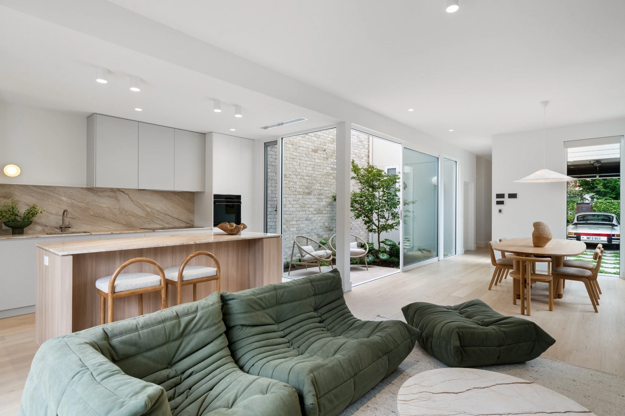 Open-concept living room and kitchen with minimalist design, large windows, indoor plants, and wooden furniture.