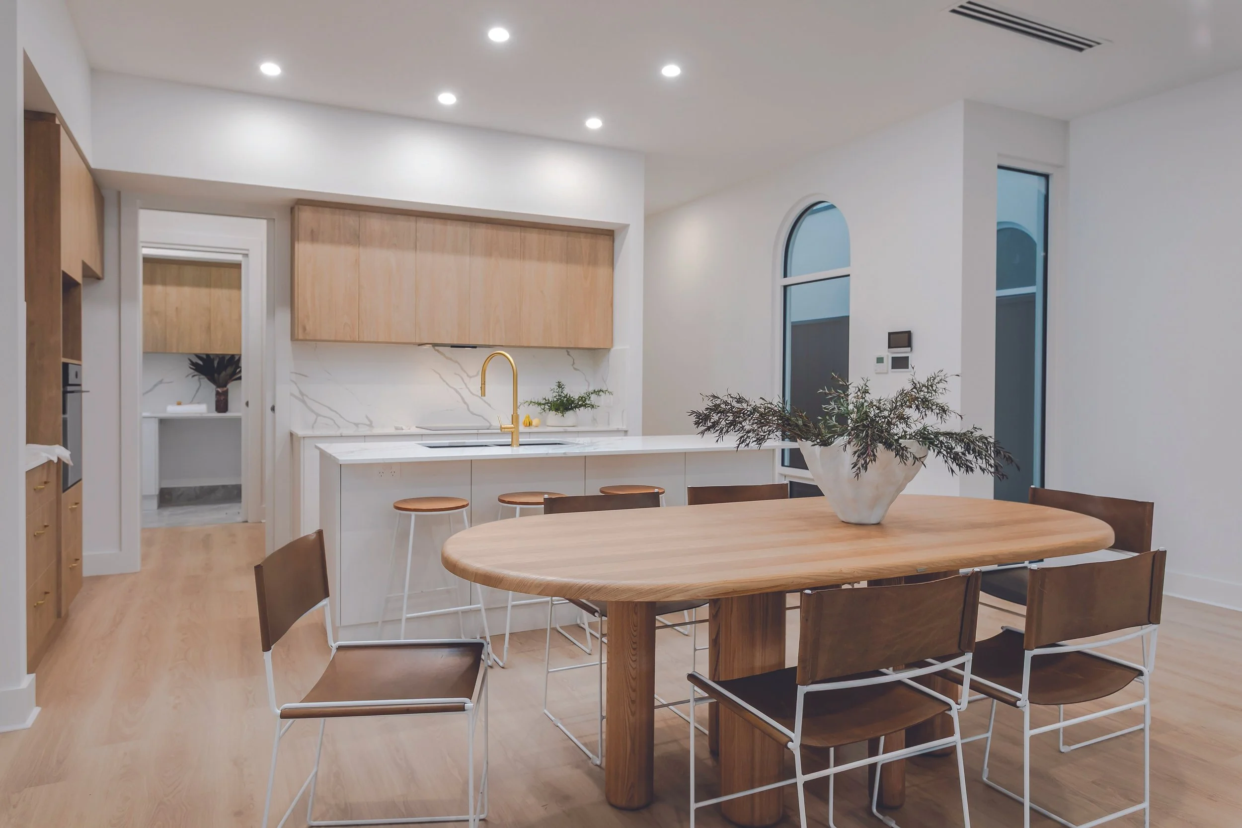 Modern kitchen and dining area with wooden and white accents, a round wooden dining table with matching chairs, a kitchen island with a white marble countertop, a gold faucet, and decor including a cactus plant in a white pot.