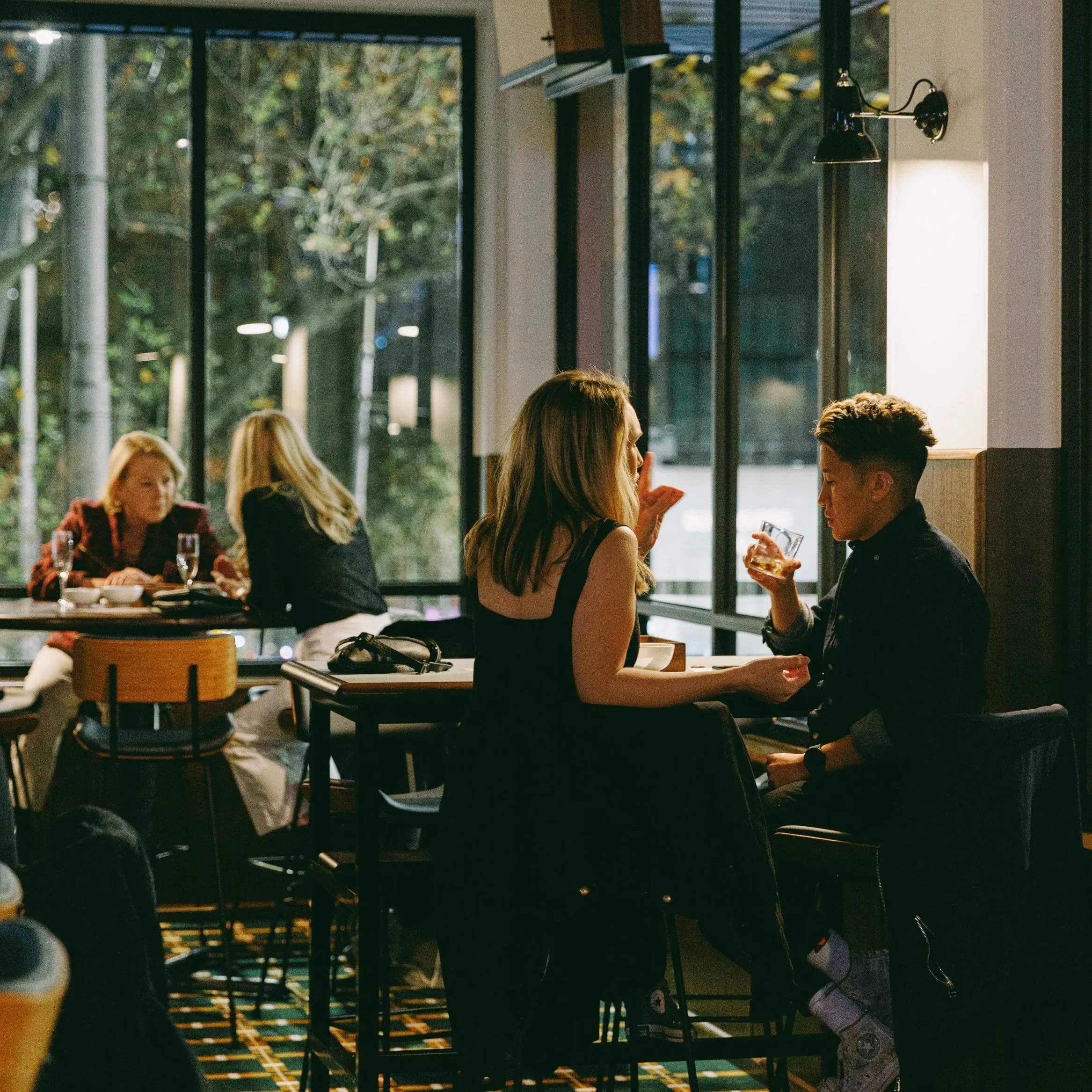 People sitting at tables inside a restaurant with large windows showing trees outside. Some are engaged in conversation, and one is holding a drink.