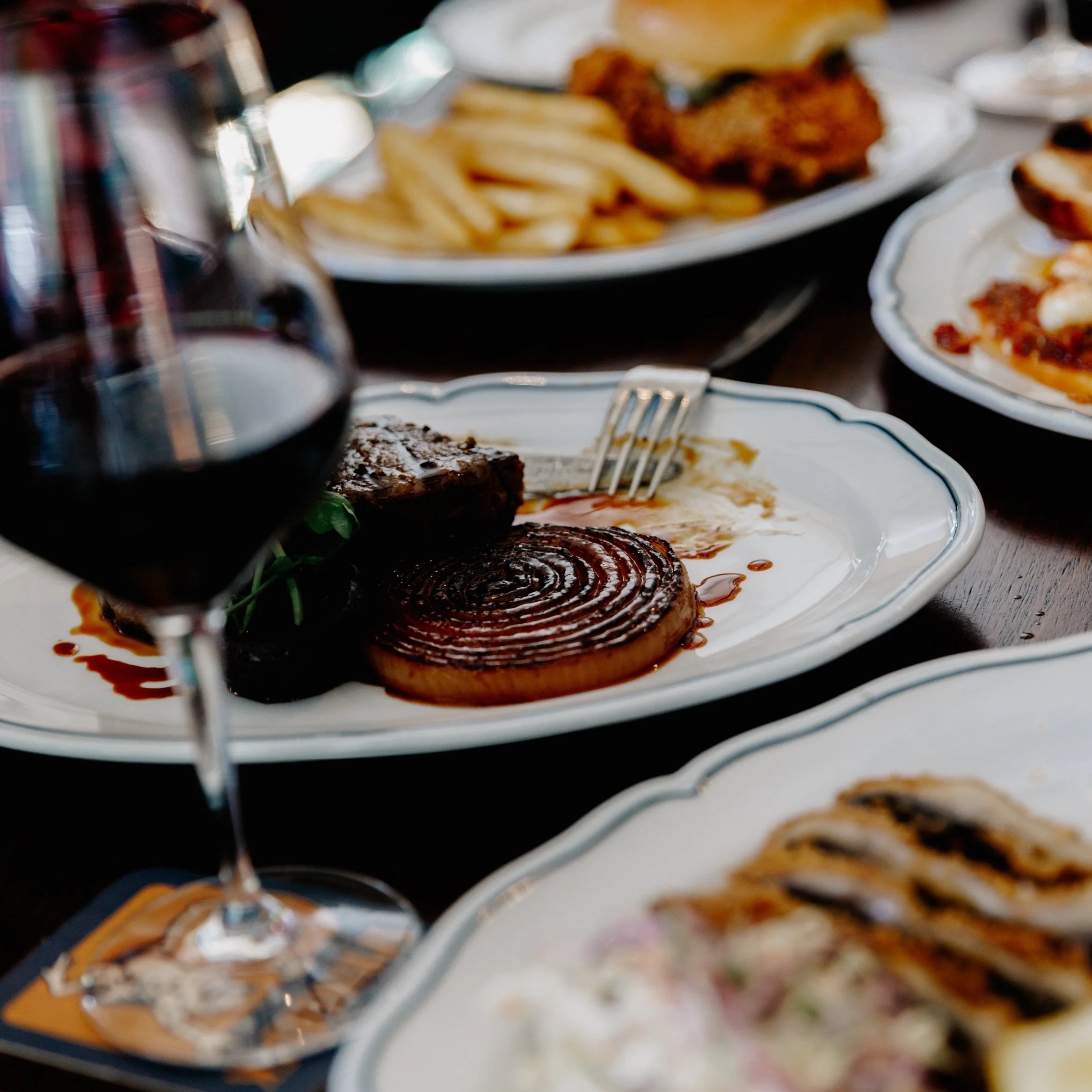Close-up of a restaurant table with plates of food, including a grilled steak, French fries, fried chicken, and dishes with sauce, accompanied by a glass of red wine.