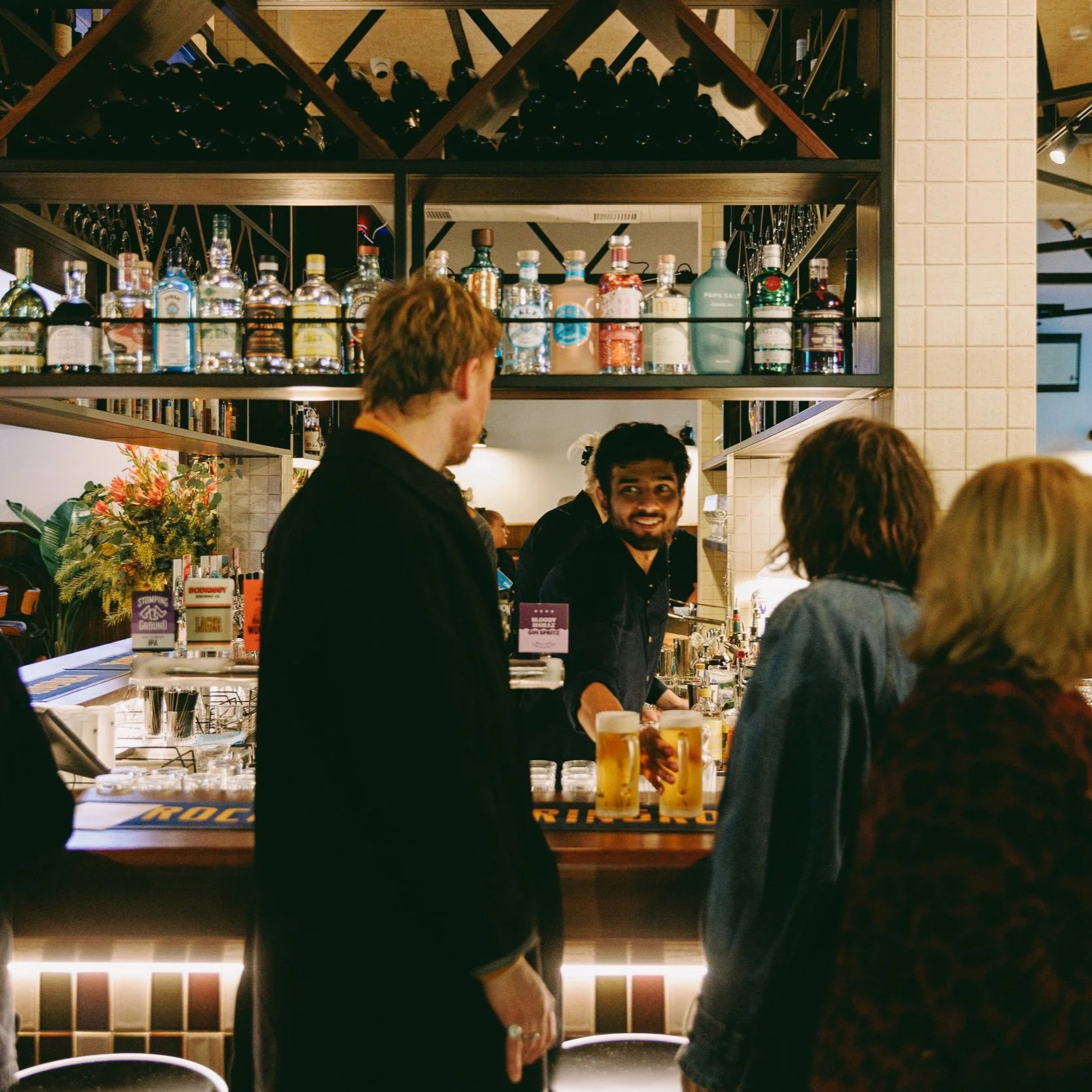 People at a bar counter with drinks, bar shelves with bottles of alcohol, and bar staff serving customers in a restaurant or pub setting.