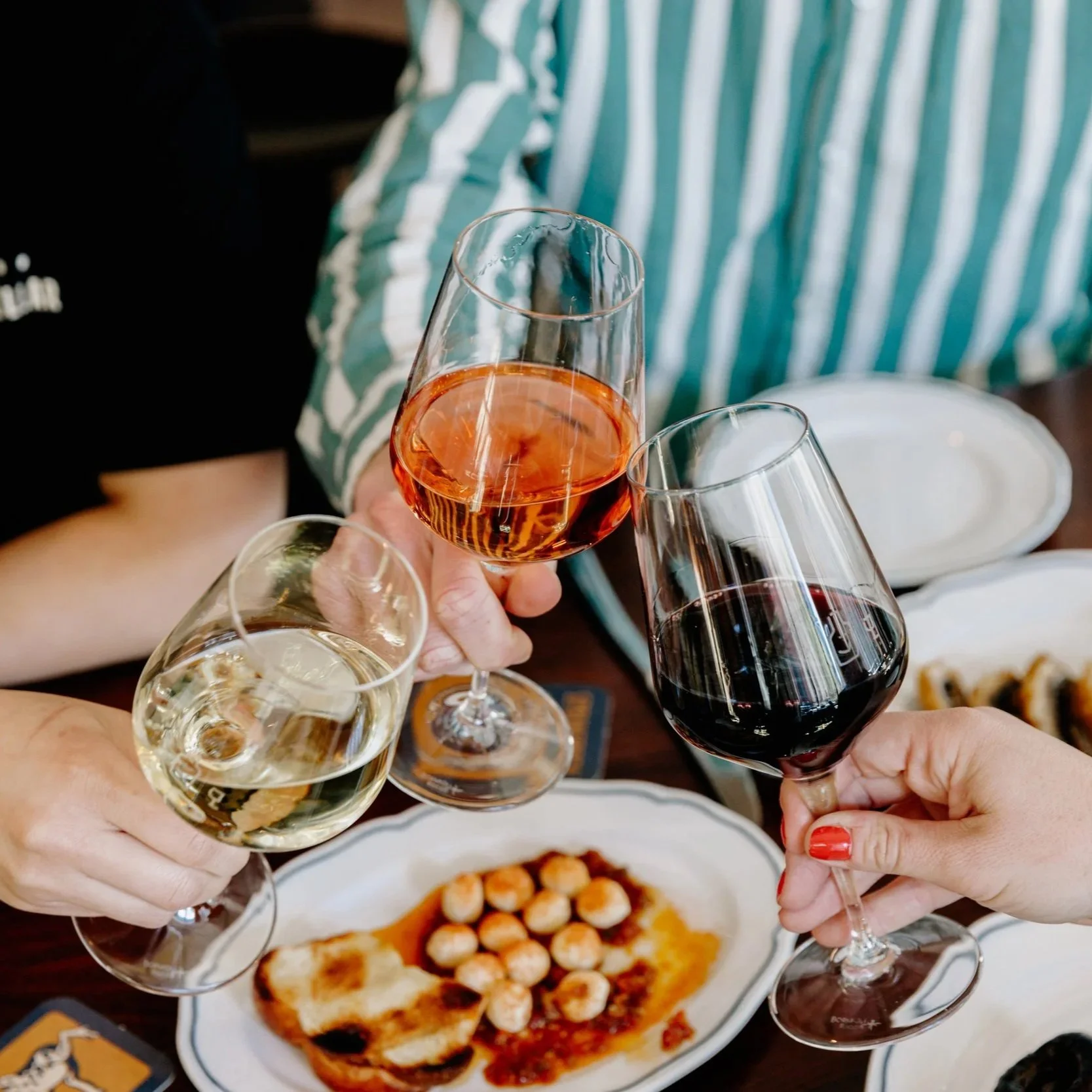 People clinking glasses of white wine, rosé, and red wine in celebration at a dinner table with a plate of garlic bread and a dish topped with small round appetizers.
