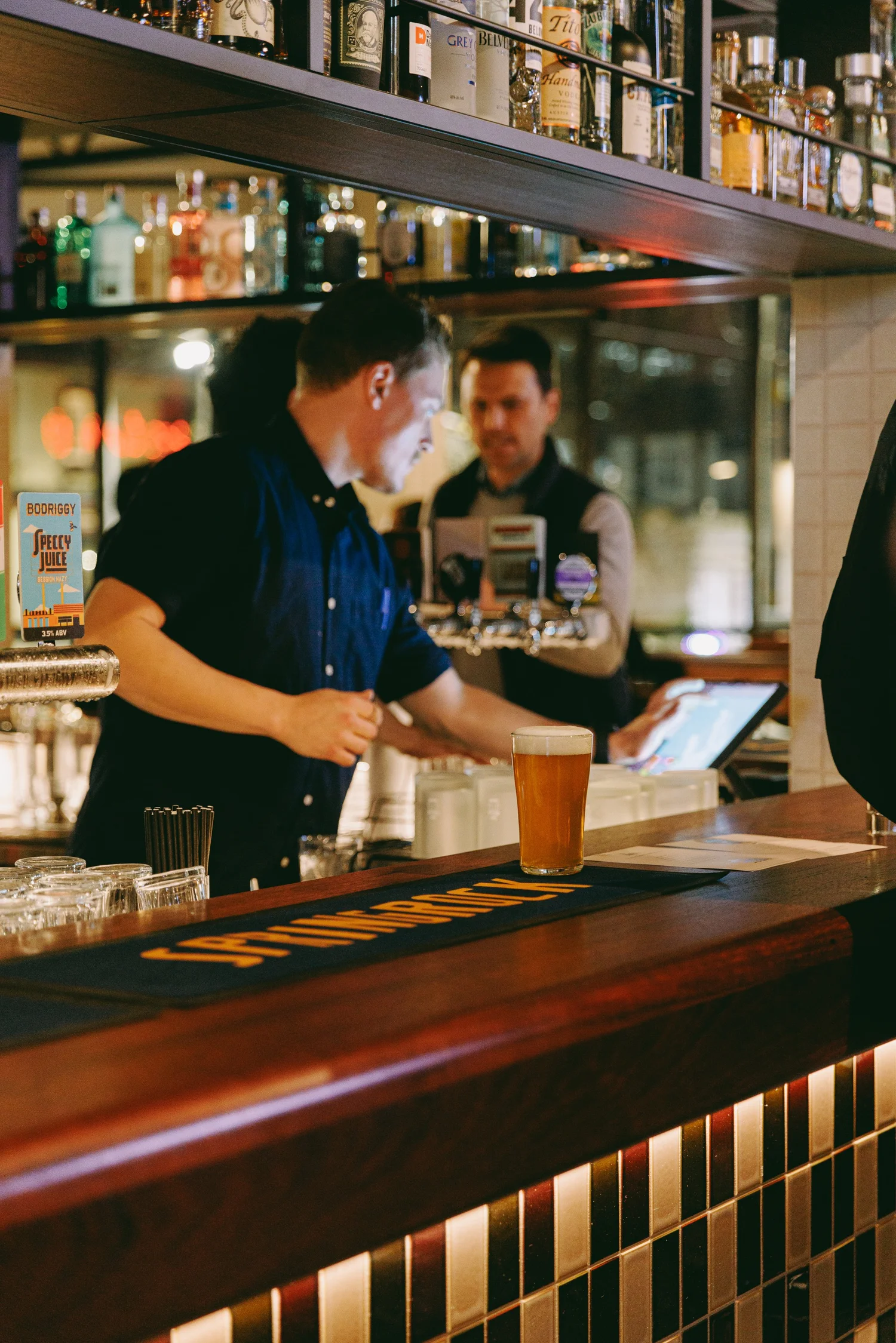 Two bartenders working behind the Springrock bar, with pints poured and bottles lining the shelves