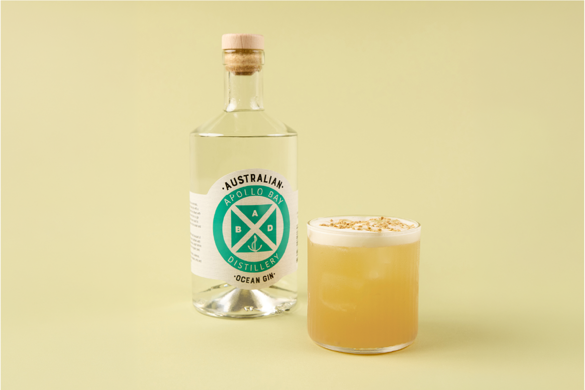 A clear glass bottle with a cork top labeled 'Australian Apollo Bay Distillery Ocean Gin' next to a glass of light yellow gin with a foamy top and sprinkled herbs or botanicals.