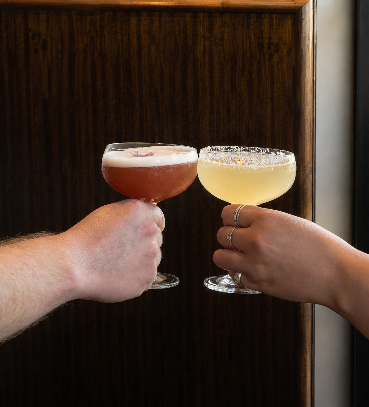 Two people clinking glasses of cocktails, one reddish and one yellow, against a dark wooden background.