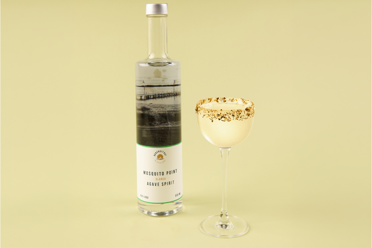 A bottle of Mosquito Point Blanco Agave Spirit and a cocktail glass with a salted rim on a light yellow background.